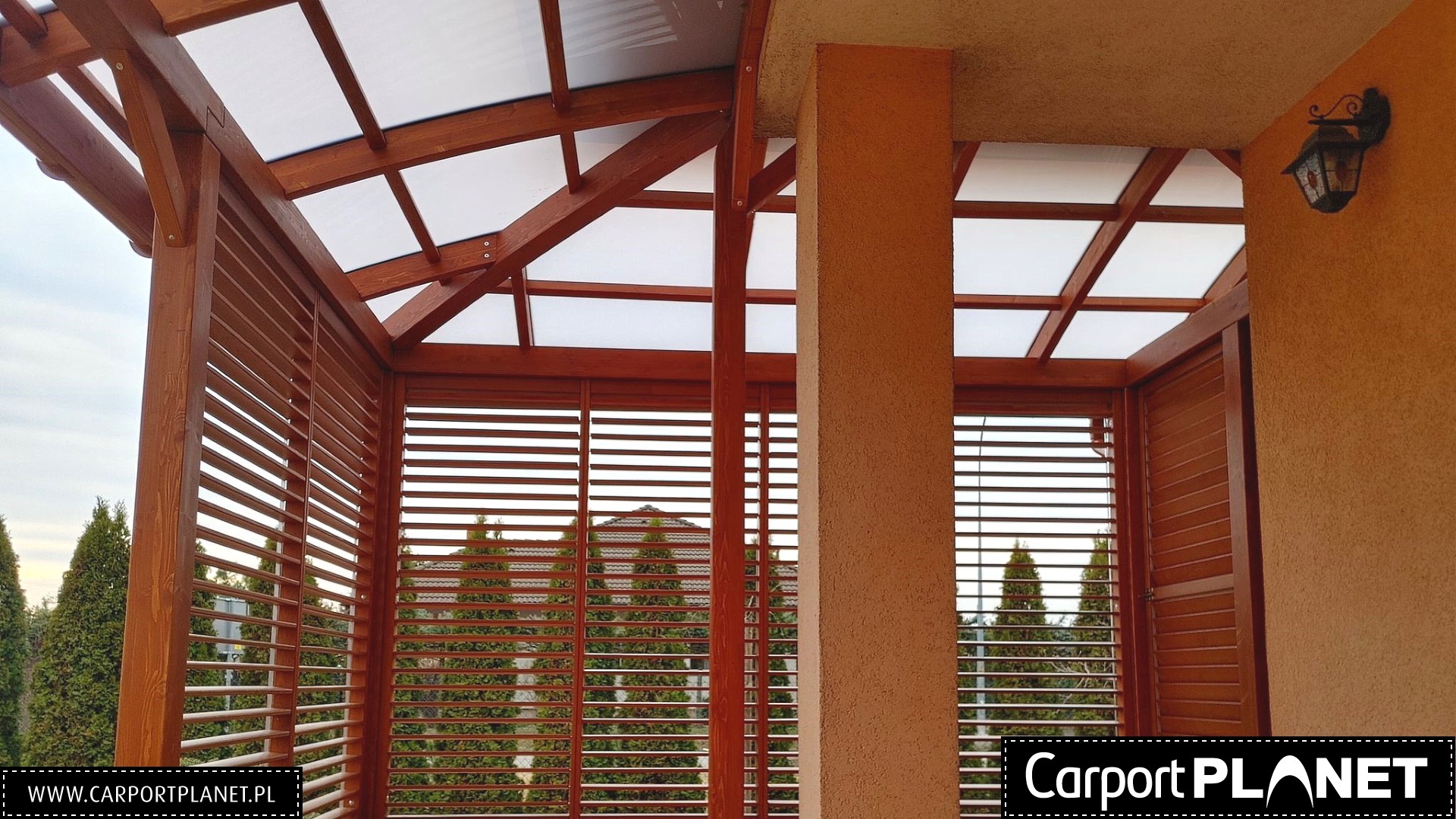 Drewniana konstrukcja z lamelami i przezroczystym zadaszeniem, widok z wnętrza na zewnątrz, w tle zieleń i zabudowania.