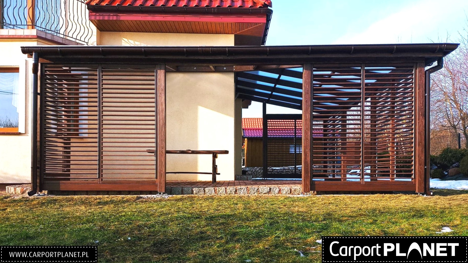 Drewniana konstrukcja z lamelami, prawdopodobnie zadaszenie tarasu lub altana, z widocznym fragmentem budynku mieszkalnego w tle i elementami ogrodu.