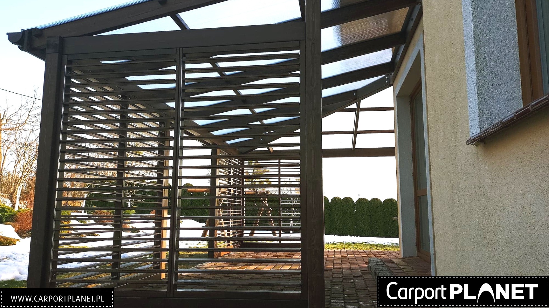 Zadaszona konstrukcja z drewnianymi lamelami, przylegająca do budynku, z widocznym fragmentem ogrodu pokrytego śniegiem.