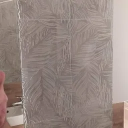 Ściana wyłożona szarymi płytkami z wytłoczonym wzorem liści, odbicie umywalki i baterii w lustrze, fragment drewnianej zabudowy.