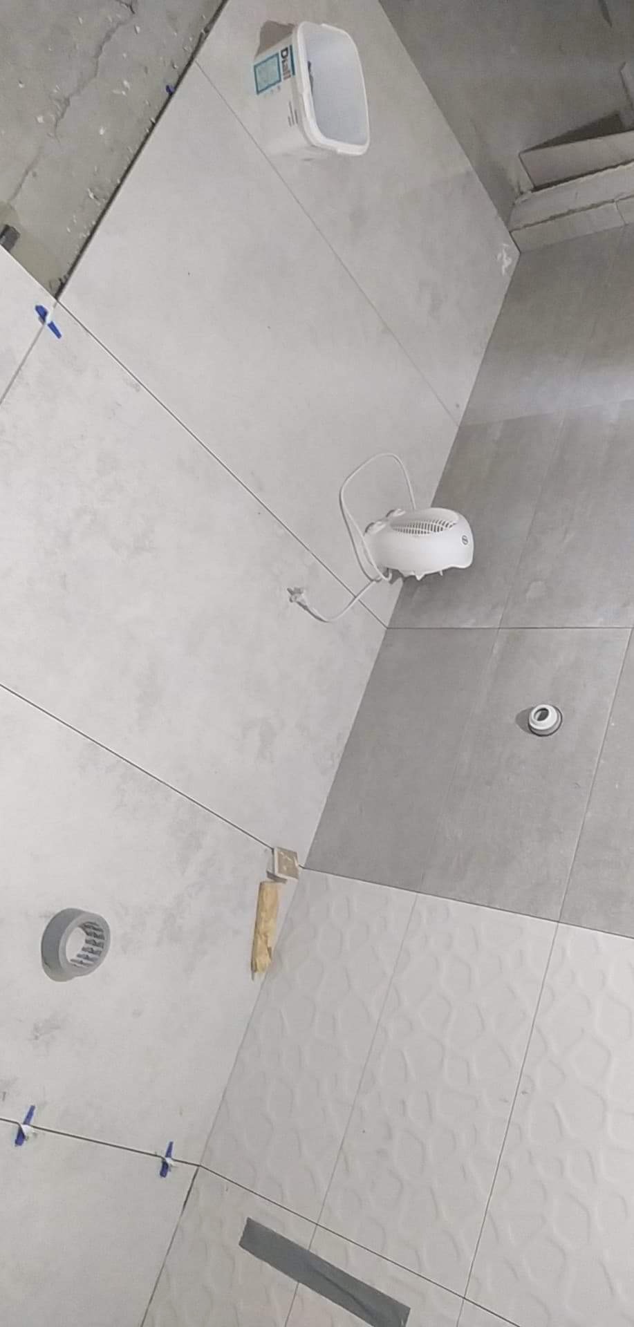 Świeżo ułożone, jasnoszare płytki w łazience, z widocznymi krzyżykami dystansowymi, poziomnicą, paskiem taśmy malarskiej, wiaderkiem, oraz małym grzejnikiem przenośnym, sugerujące trwający proces...