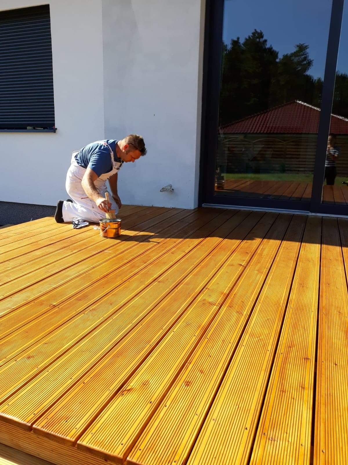 Mężczyzna w białym kombinezonie maluje drewniany taras pędzlem, widoczna puszka z bejcą, nowoczesny dom z dużymi oknami w tle.
