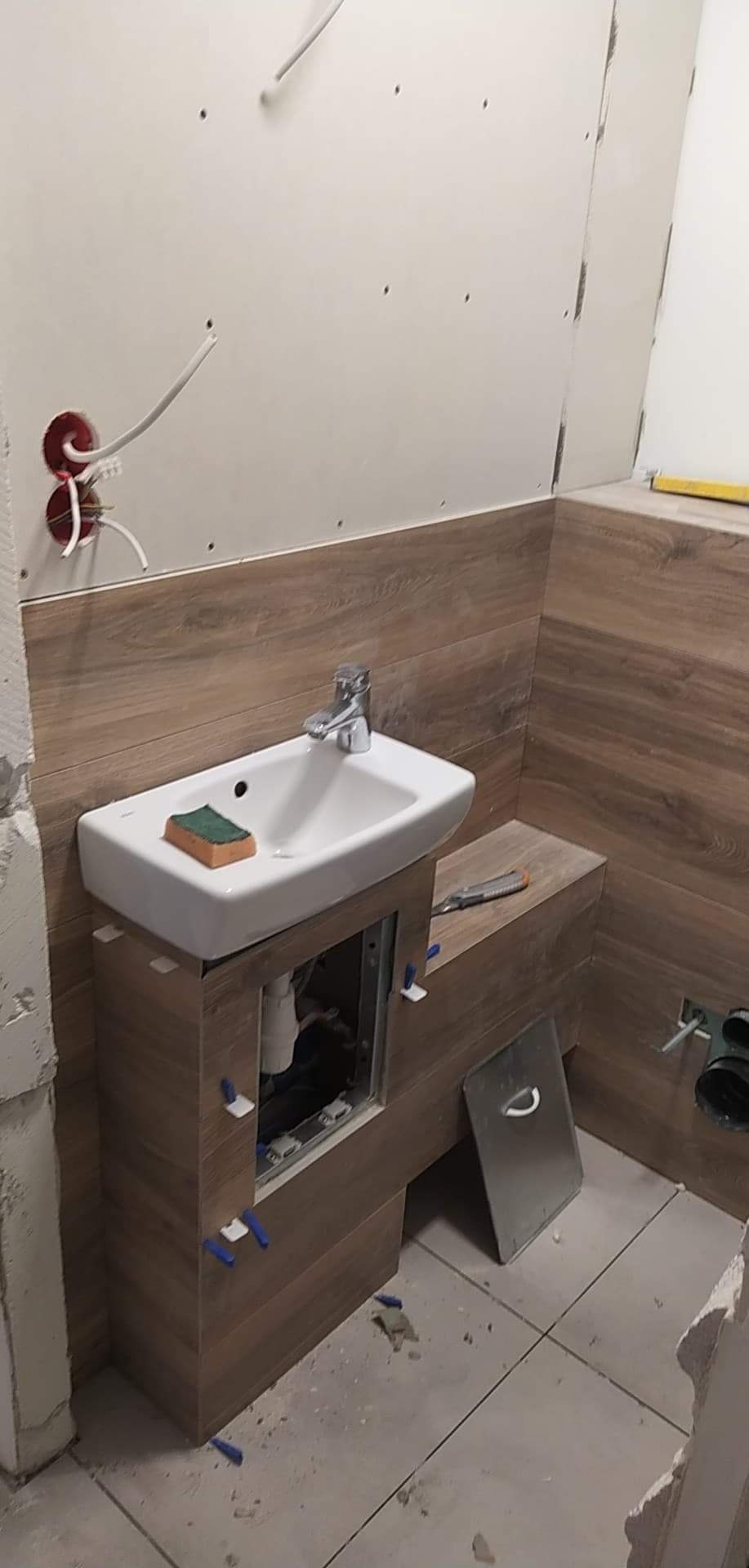 Montaż umywalki z szafką w łazience w trakcie prac wykończeniowych, widoczne płytki imitujące drewno na ścianie, biała umywalka z baterią, gąbka na umywalce, narzędzia glazurnicze, niezakończona...