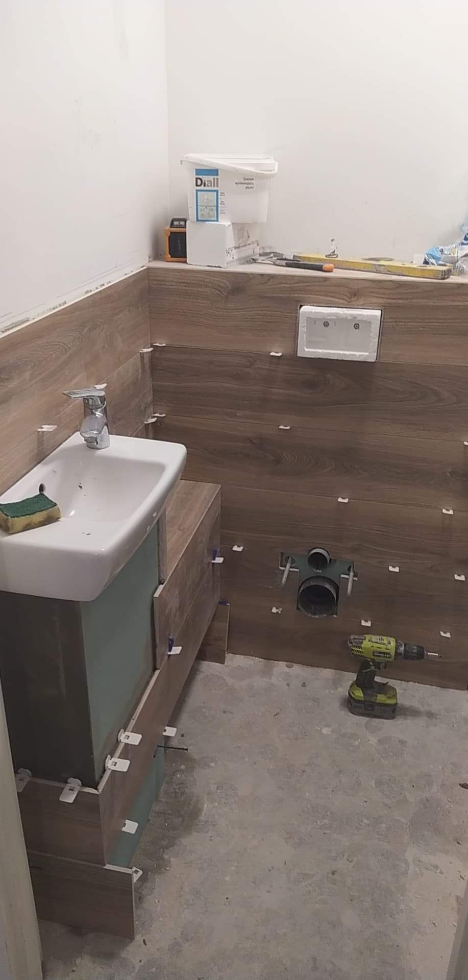 Łazienka w trakcie układania płytek imitujących drewno na ścianach, widoczna umywalka z szafką w trakcie montażu, biała spłuczka podtynkowa, narzędzia i materiały budowlane na podłodze, plastikowe...