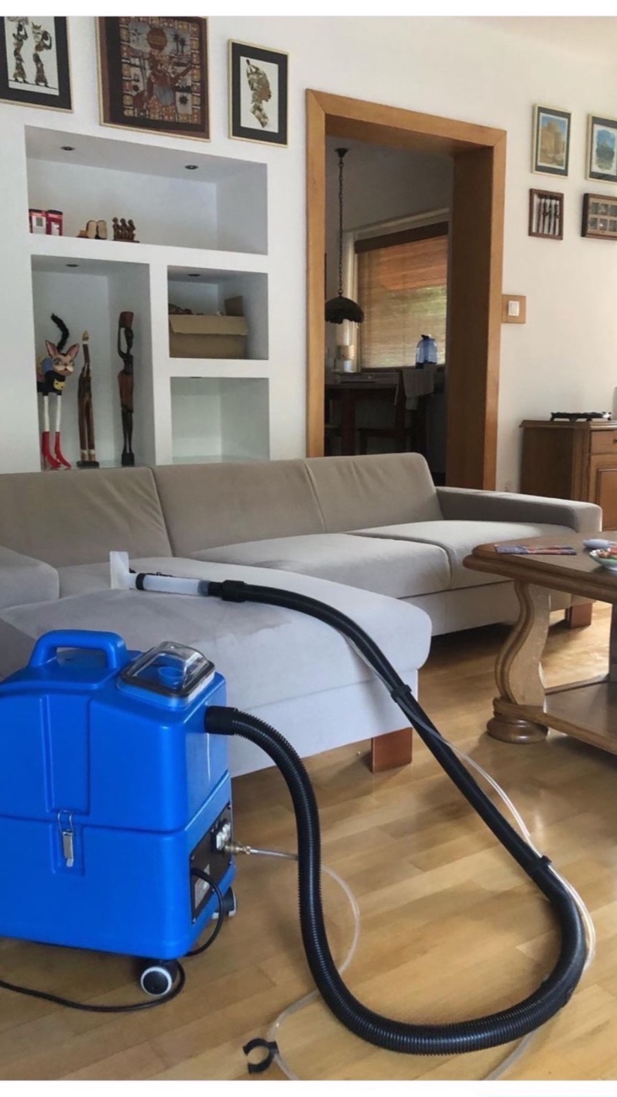 Czyszczenie jasnej kanapy za pomocą niebieskiego odkurzacza piorącego z czarnym wężem w salonie z drewnianą podłogą i dekoracjami na ścianach.