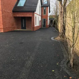 Nowoczesny dom z czerwonej cegły z białymi akcentami, widok z zewnątrz na świeżo położony, czarny asfaltowy podjazd z obrzeżem z kostki brukowej, drzewo z gołymi gałęziami przy murze z cegły.