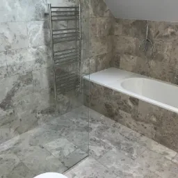 Wykończona łazienka z szarymi marmurkowymi płytkami, białą wanną i grzejnikiem drabinkowym, oddzielona szklaną ścianką prysznicową.