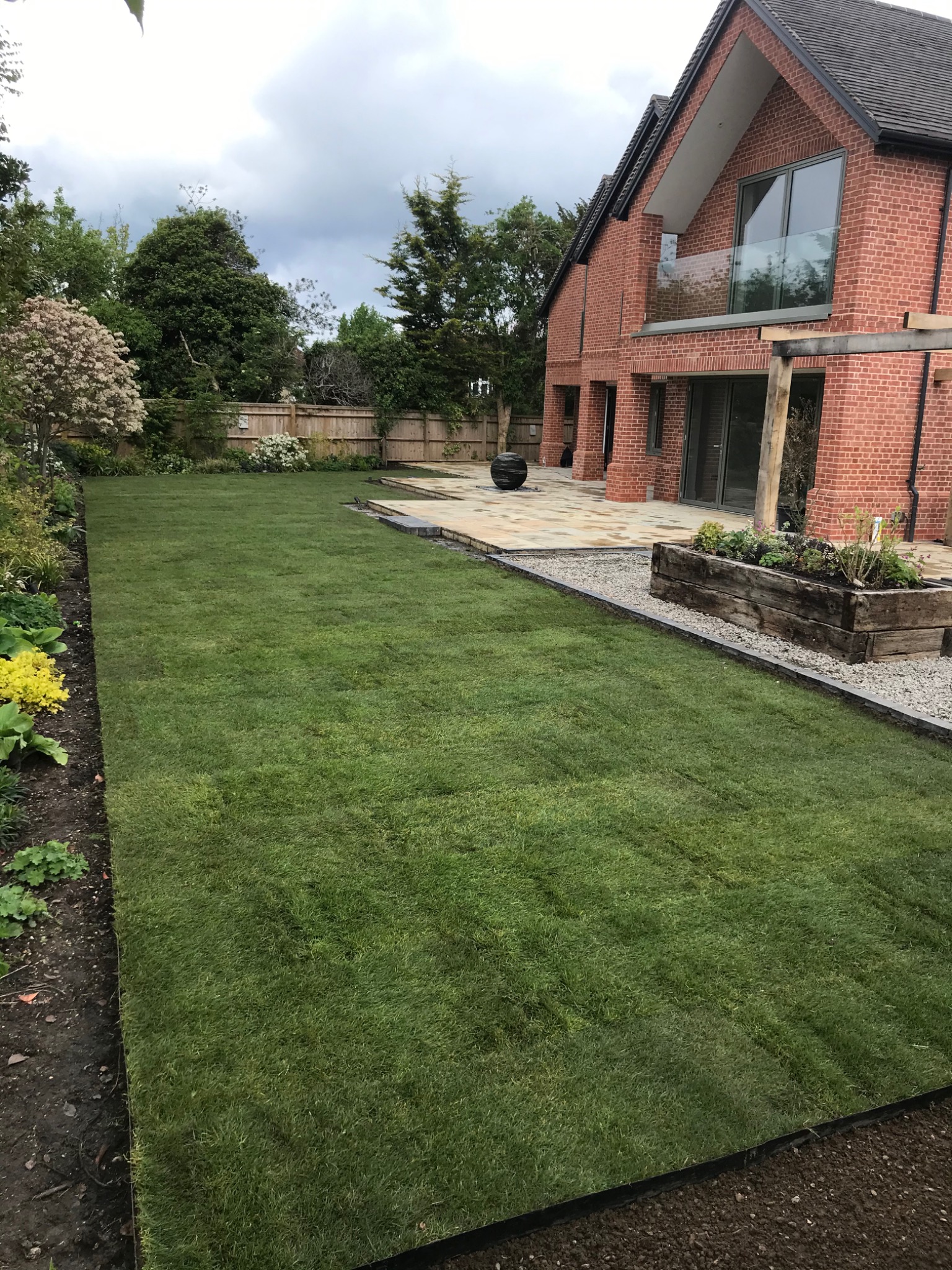 Nowo rozłożony trawnik z rolki przylegający do tarasu z piaskowca i rabaty z drewnianą skrzynią, w tle nowoczesny dom z czerwonej cegły i drewniany płot.