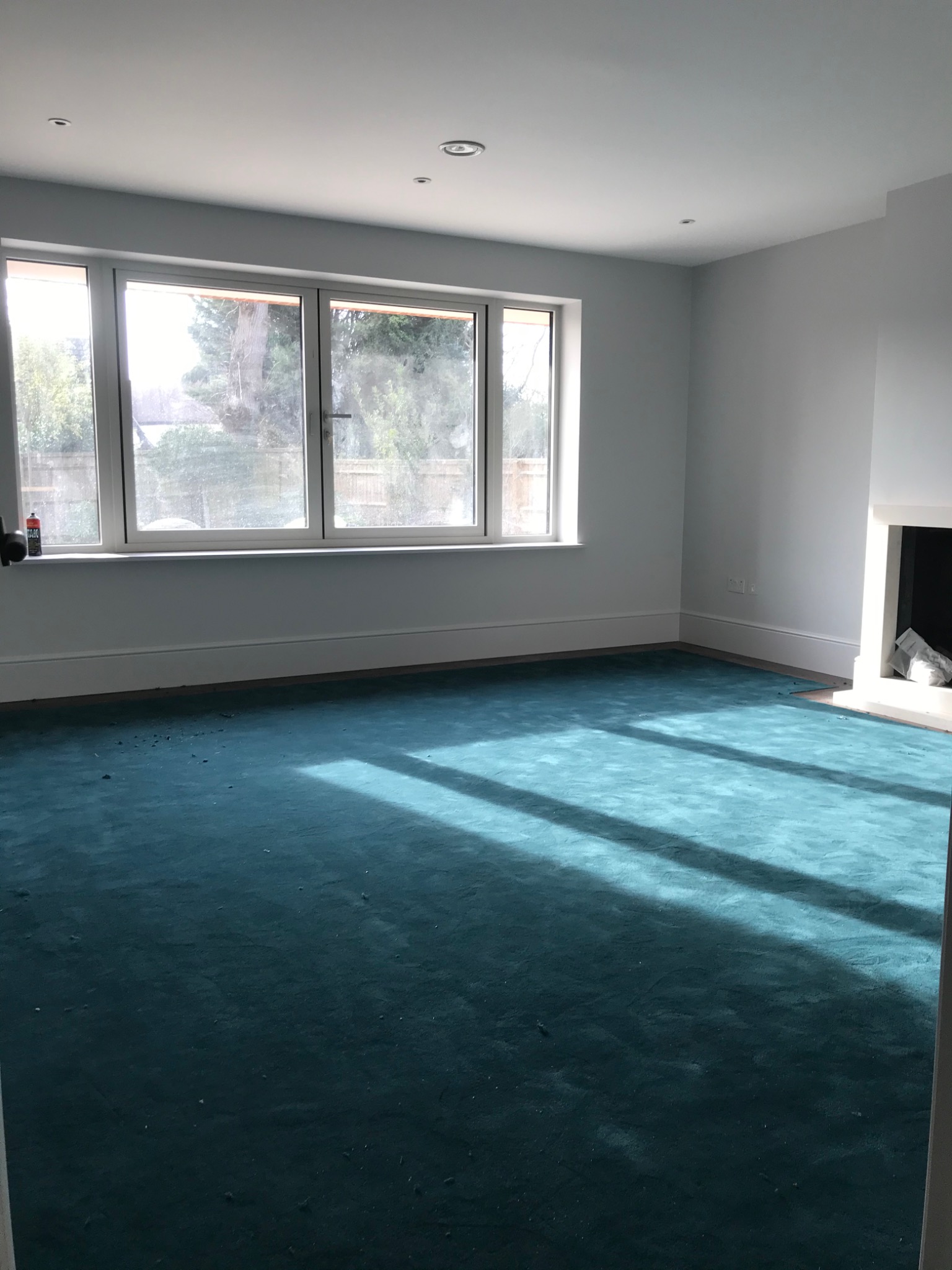 Pusta, jasna przestrzeń z turkusową wykładziną, dużym oknem z widokiem na zieleń i kominkiem, przygotowana do dalszego wykończenia.
