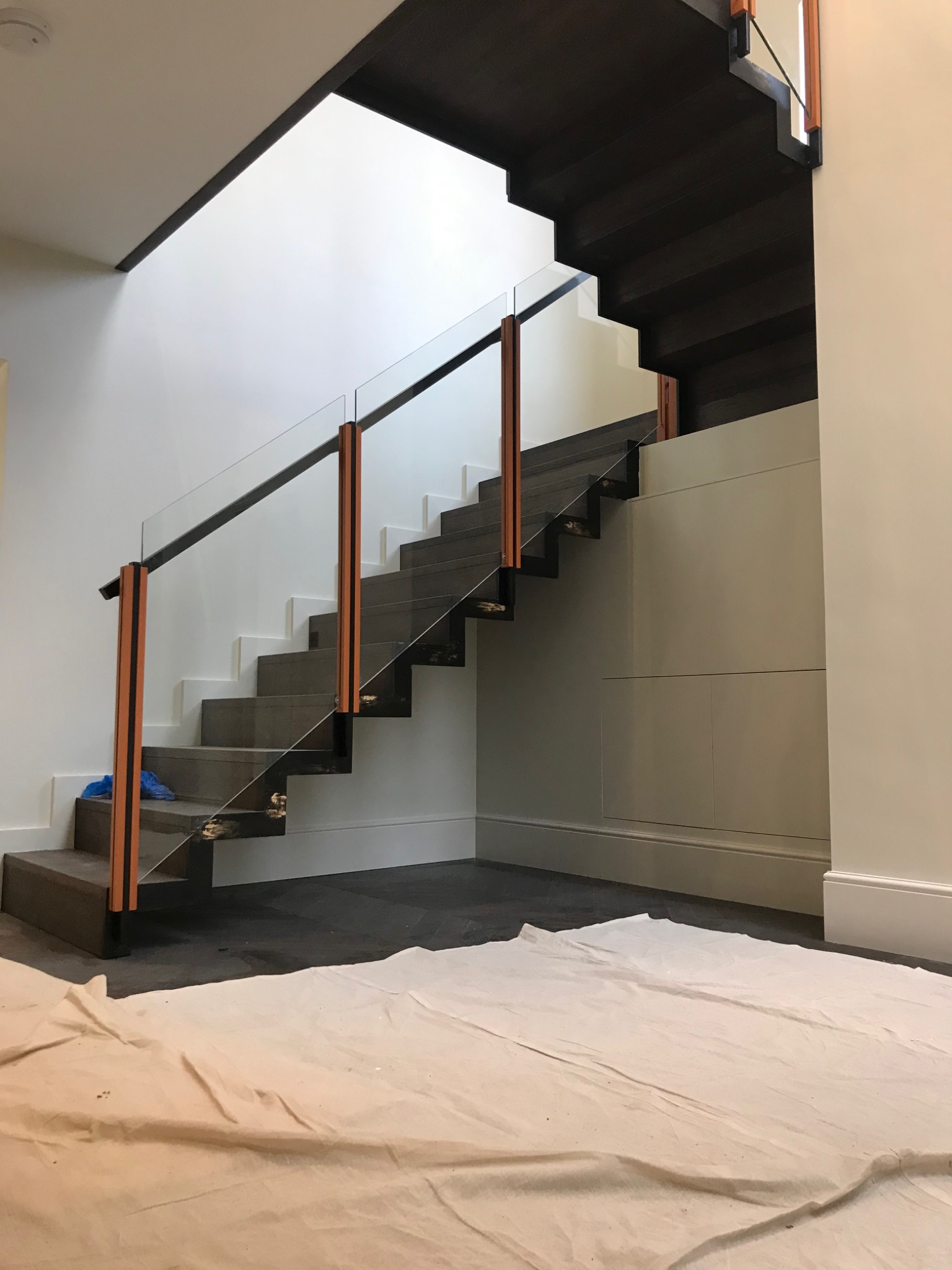 Nowoczesne schody z ciemnego drewna i szklanymi balustradami z drewnianymi słupkami, zabezpieczone płachtą malarską na podłodze, widok z dołu.