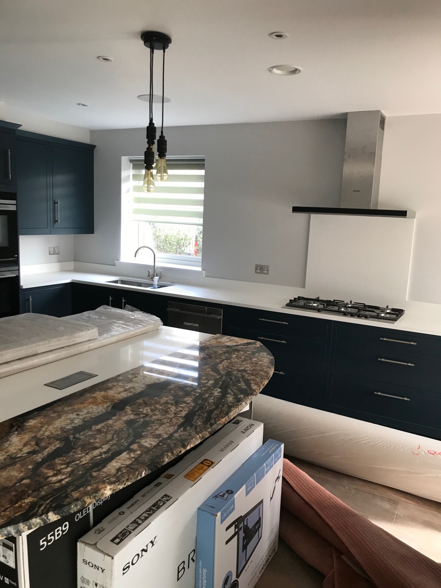 Nowoczesna kuchnia z granatowymi szafkami, białym blatem i wyspą z blatem imitującym kamień. Na blacie kuchennym płyta gazowa i okap. Na wyspie leżą zapakowane elementy, w tym kartony z logo Sony.