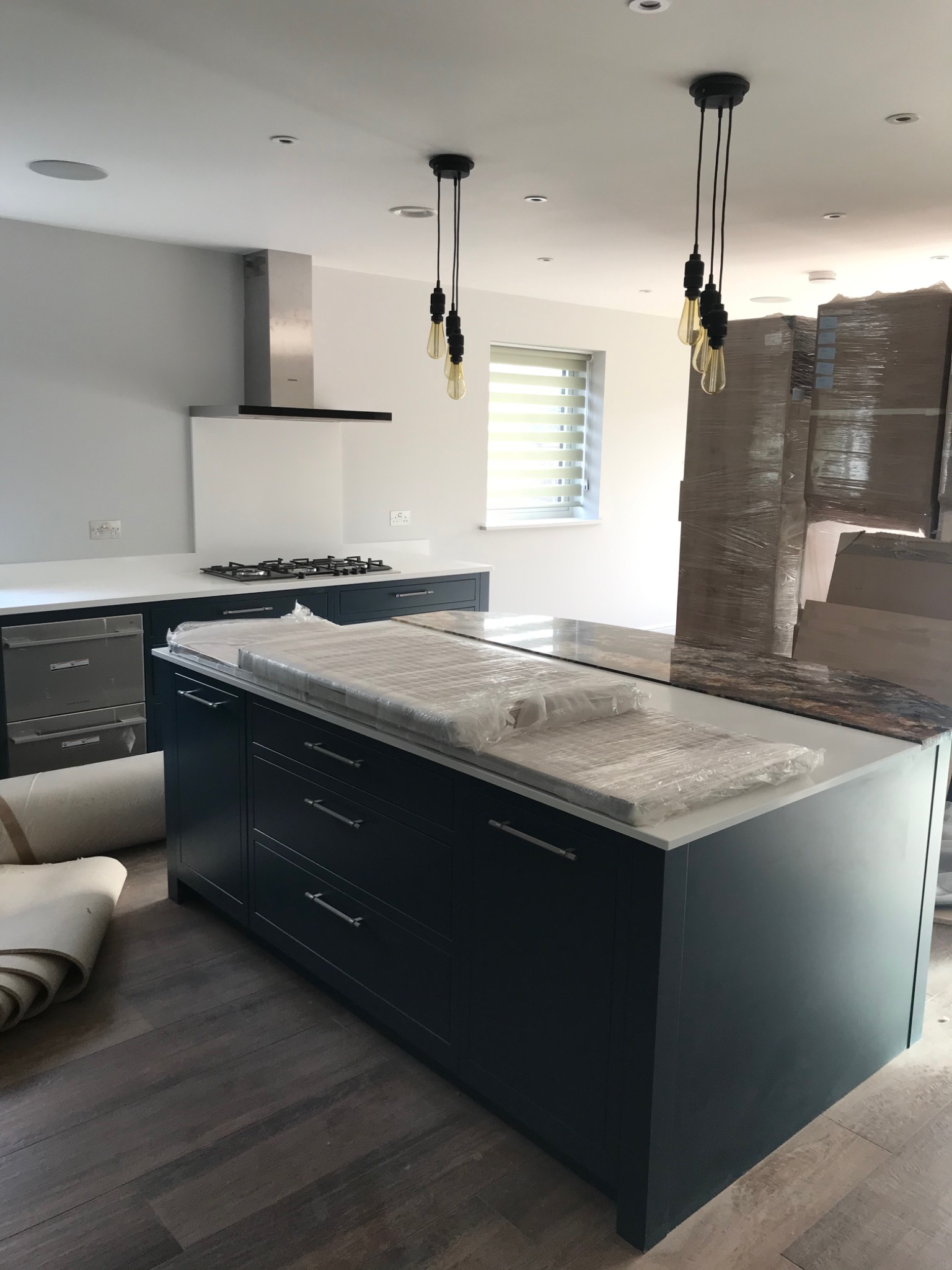 Nowoczesna kuchnia w trakcie wykończenia: ciemnogranatowa wyspa kuchenna z białym blatem zabezpieczonym folią, w tle sprzęt AGD, okap i rolety okienne, podłoga z paneli drewnopodobnych.