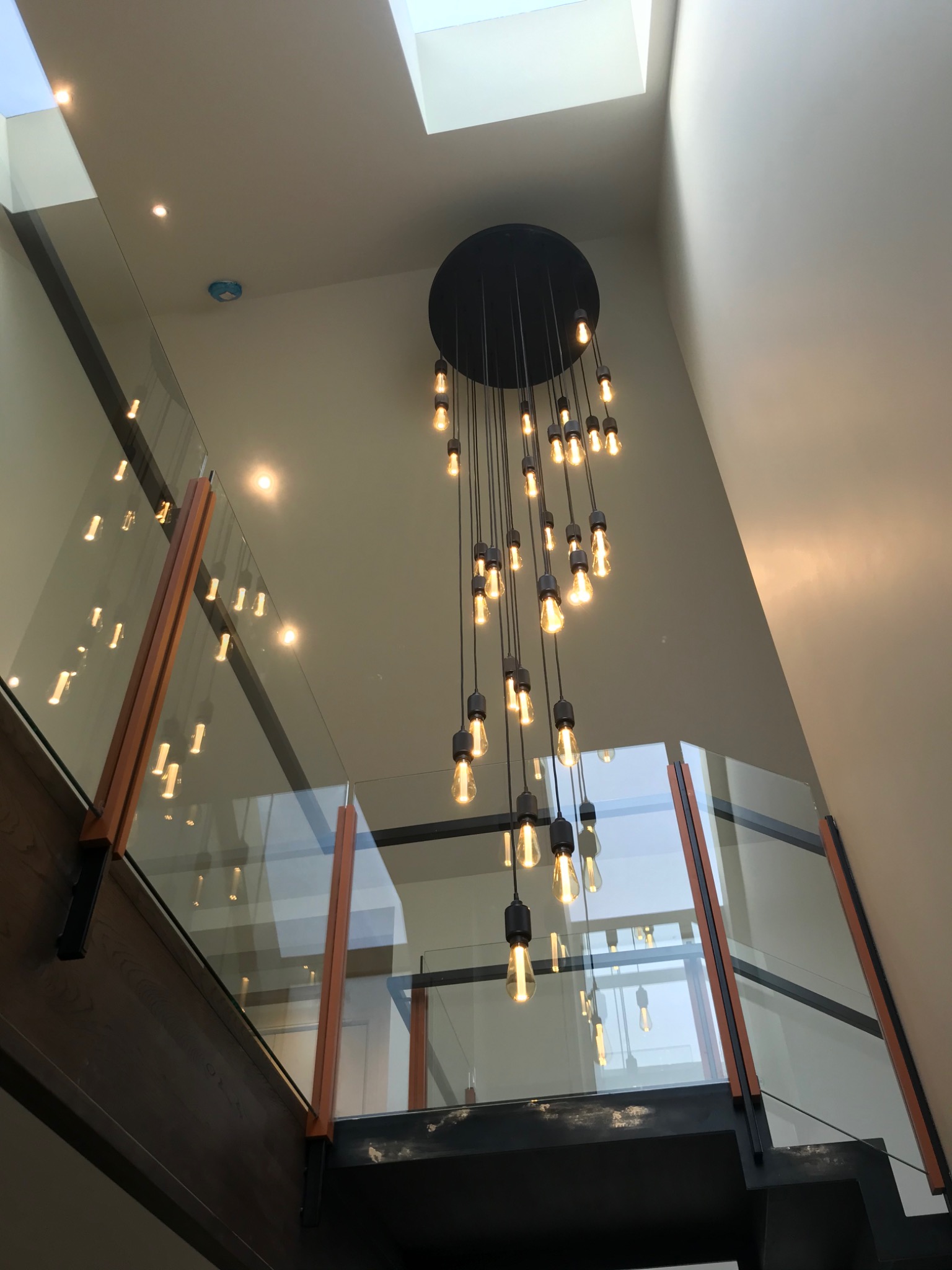 Nowoczesny żyrandol z żarówkami Edisona zwisający z sufitu w przestronnym holu z przeszkloną balustradą schodów i oknem dachowym.