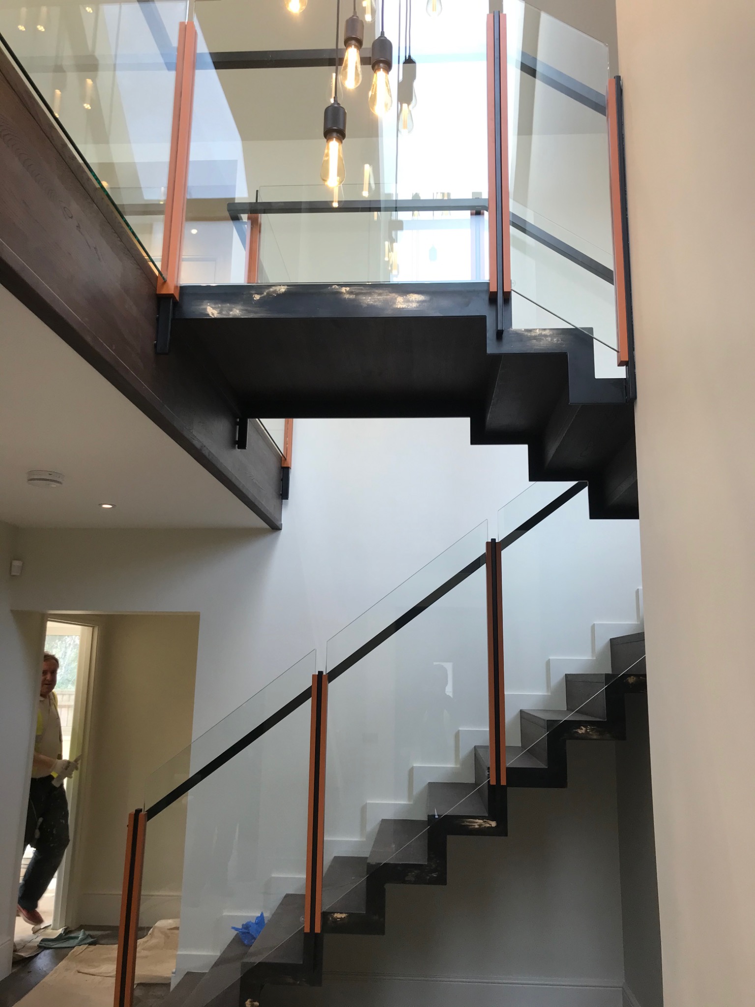 Nowoczesne schody dwubiegowe z czarnymi stopniami, białymi podstopnicami i szklanymi balustradami z pomarańczowymi akcentami, widok z dołu na antresolę z oświetleniem.