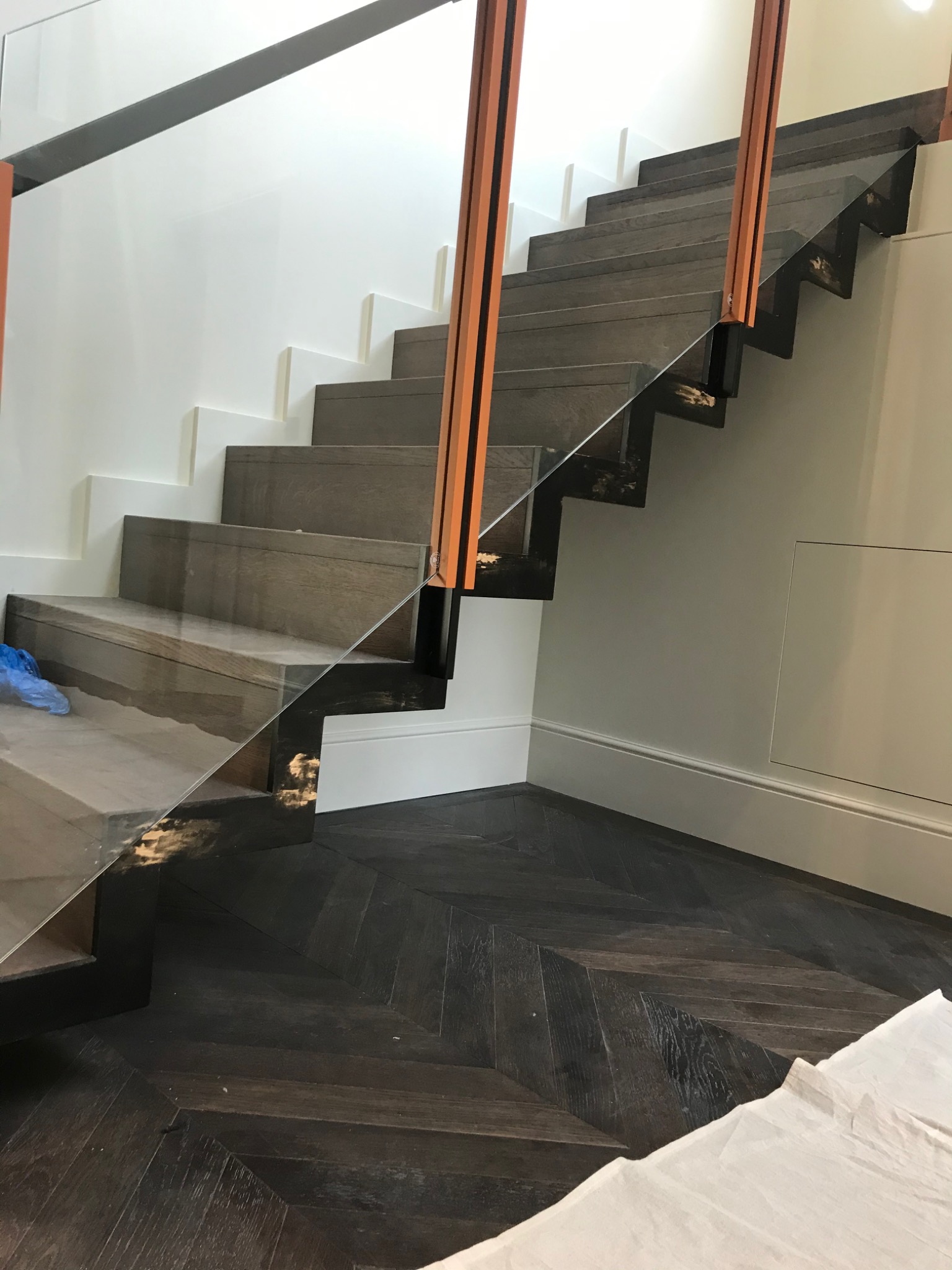 Nowoczesne schody drewniane z szklaną balustradą i podświetleniem, ułożone na ciemnej podłodze w jodełkę, widoczne zabezpieczenia na podłodze.