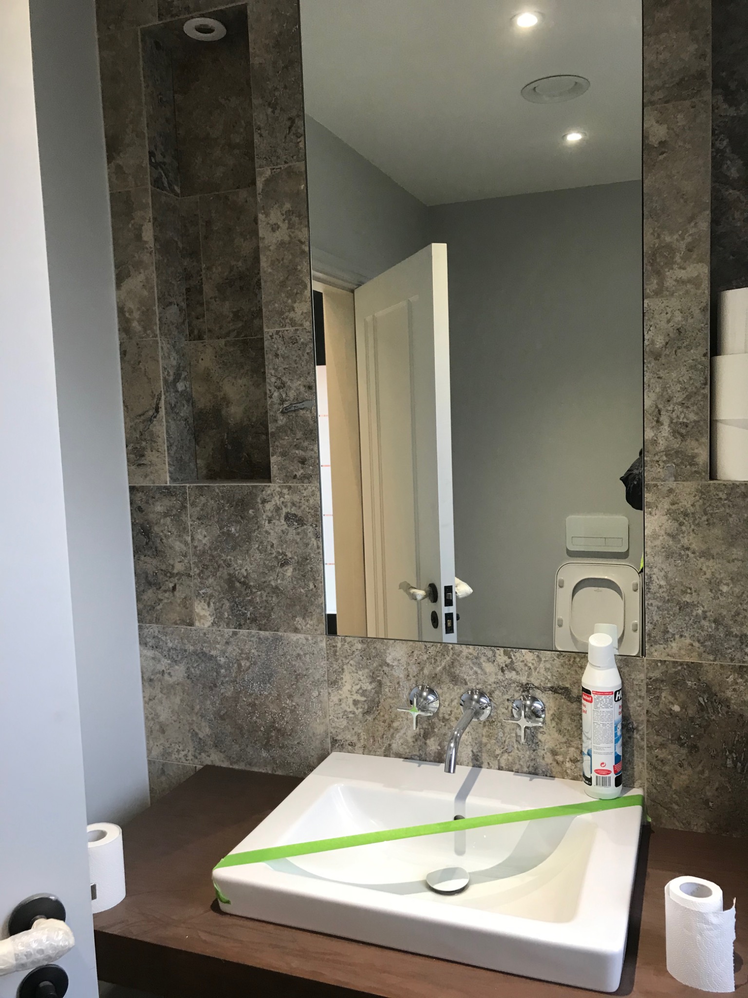 Nowoczesna łazienka z umywalką zabezpieczoną taśmą malarską, chromowaną armaturą i marmurową okładziną ścienną, widok w lustrze na otwarte drzwi i toaletę.