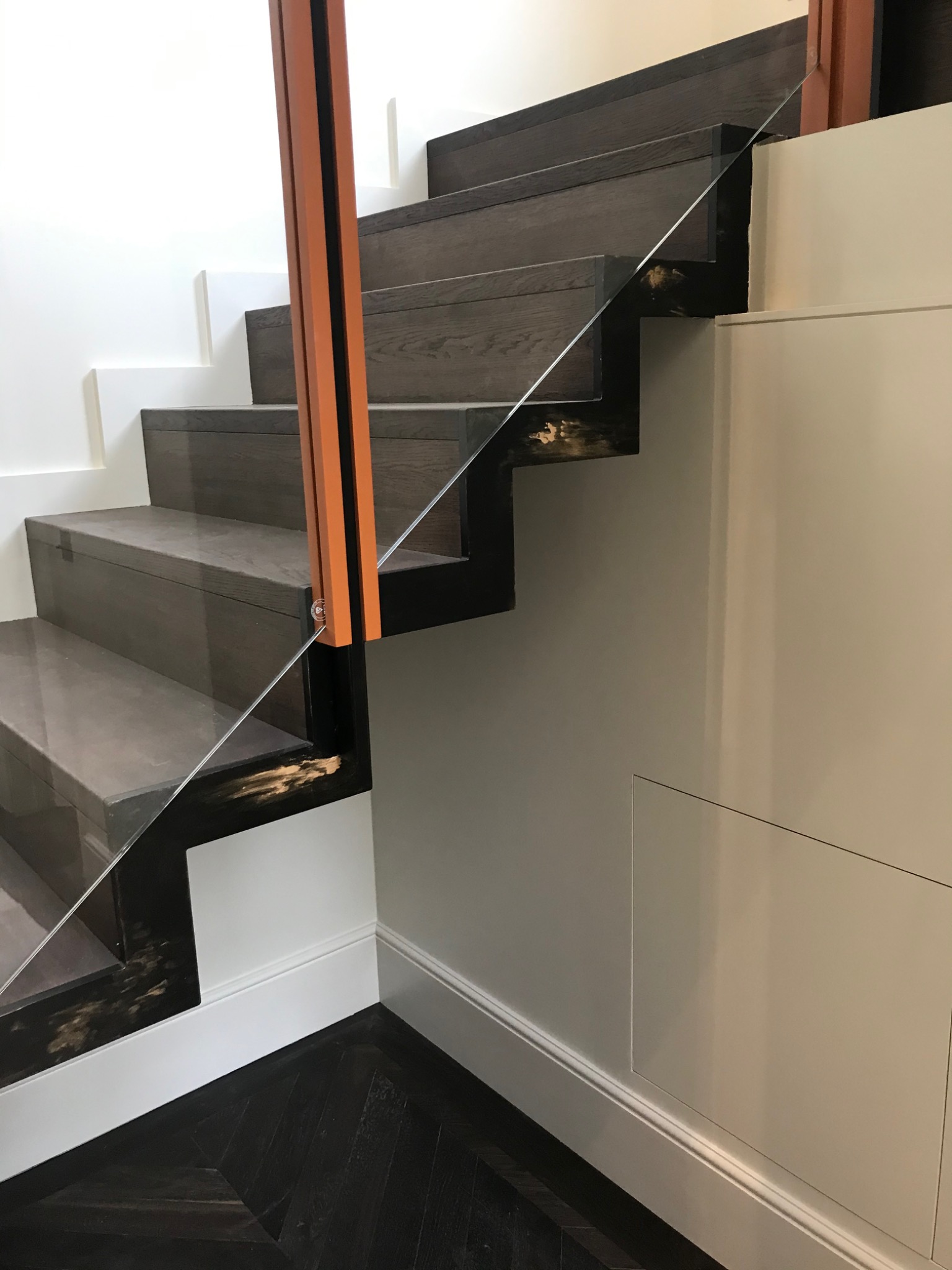 Nowoczesne schody z ciemnego drewna z szklaną balustradą i pomarańczowym słupkiem, widok z dołu na podłogę z ciemnego parkietu ułożonego w jodełkę.