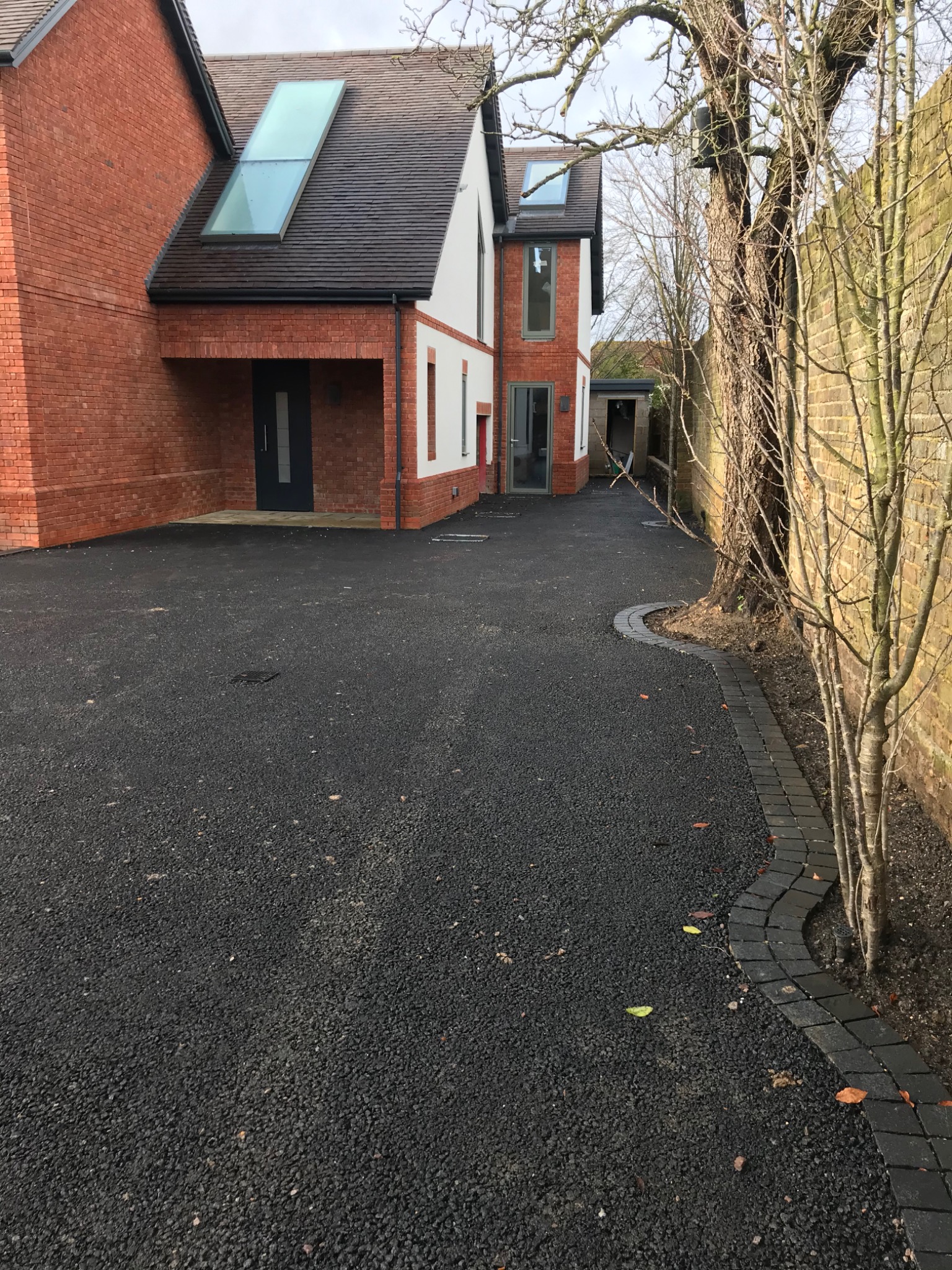 Nowoczesny dom z czerwonej cegły z białymi akcentami, widok z zewnątrz na świeżo położony, czarny asfaltowy podjazd z obrzeżem z kostki brukowej, drzewo z gołymi gałęziami przy murze z cegły.