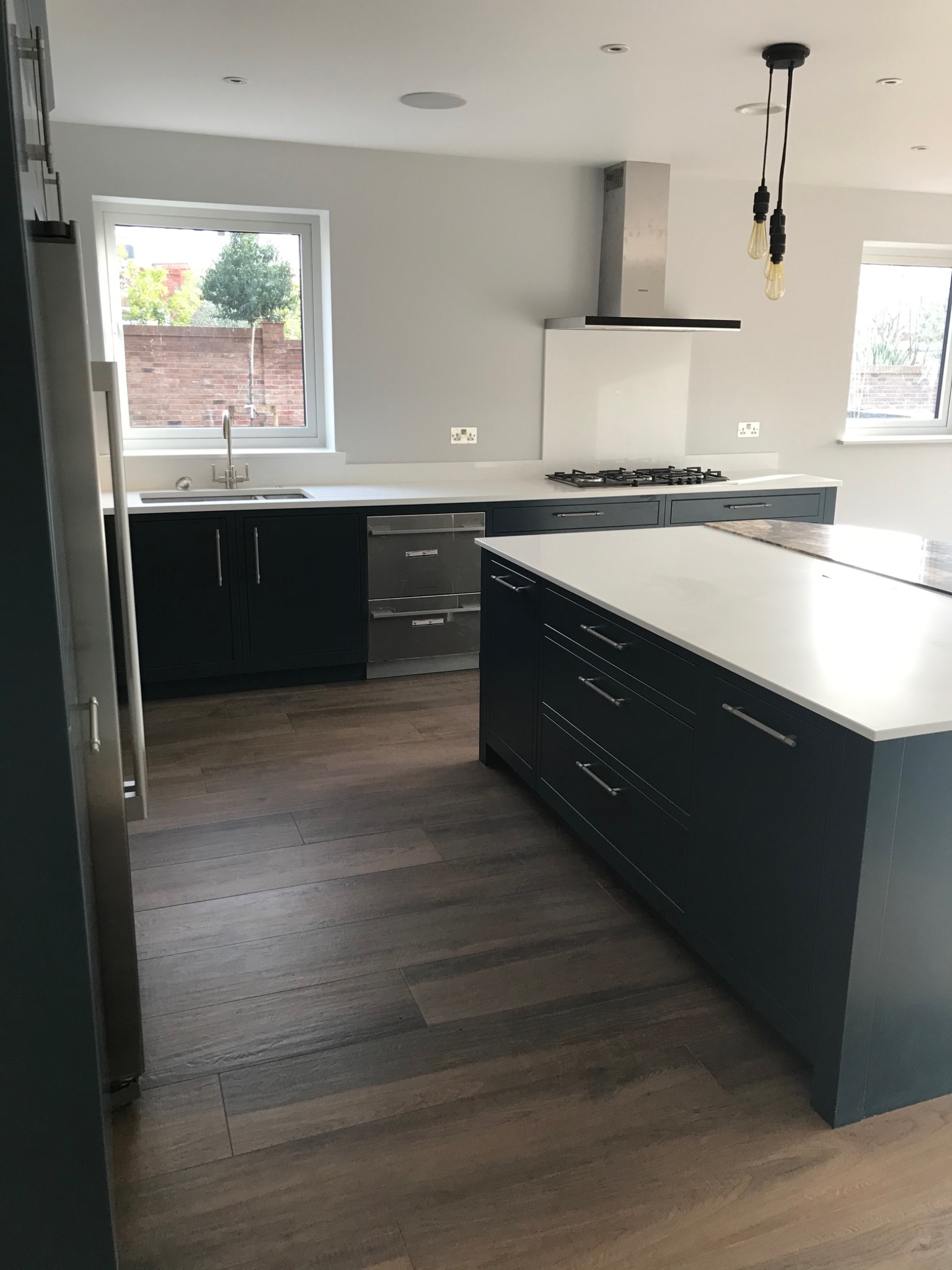 Nowoczesna kuchnia z ciemnoniebieskimi szafkami, białym blatem i drewnopodobną podłogą, widok na okno z zielenią i ceglanym murem w tle, minimalistyczny design.