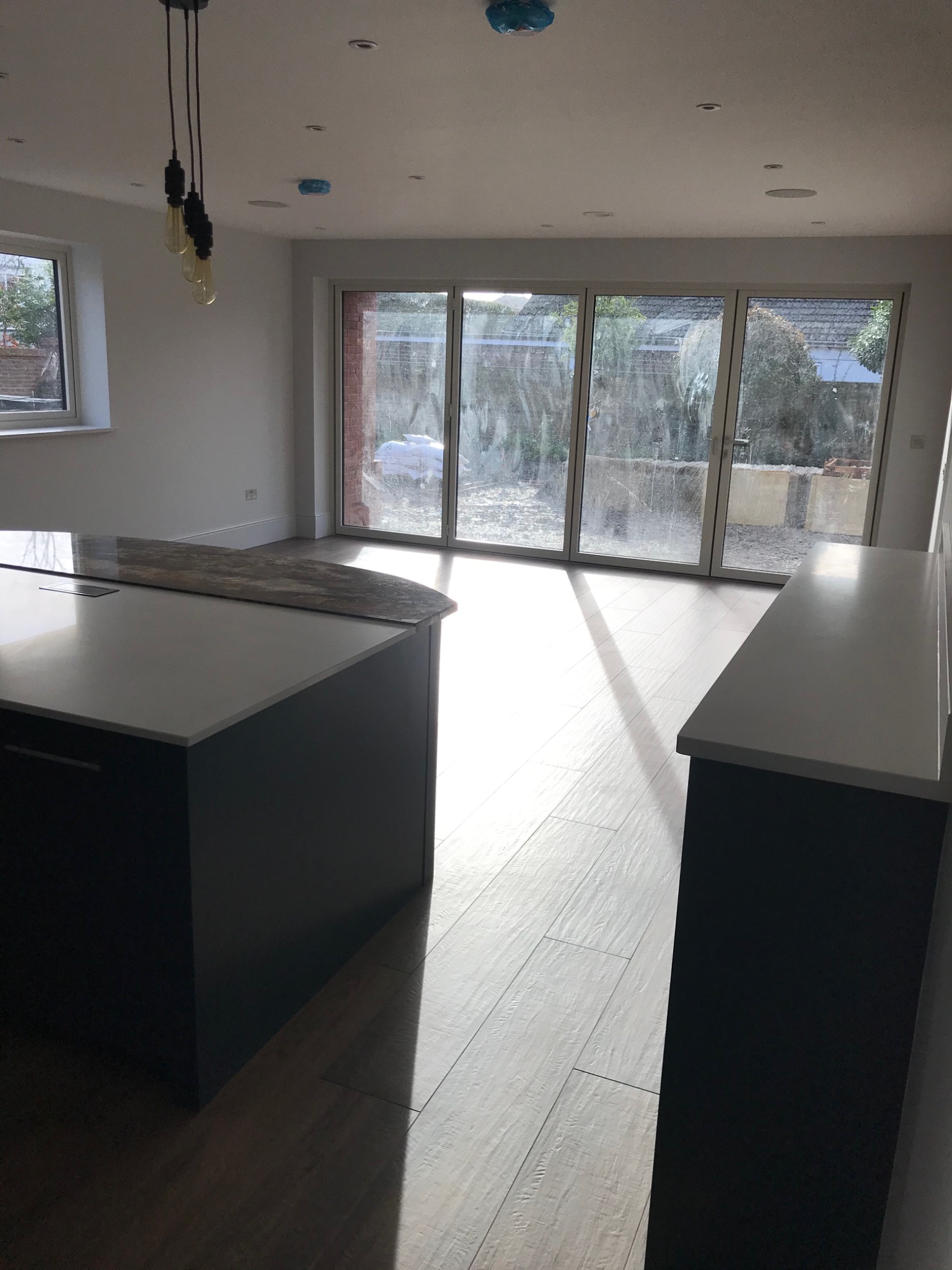 Jasne wnętrze domu z drewnianą podłogą, wyspą kuchenną z blatem z konglomeratu i dużymi przesuwnymi oknami wychodzącymi na ogród. Widoczne lampy wiszące w stylu industrialnym i elementy instalacji...