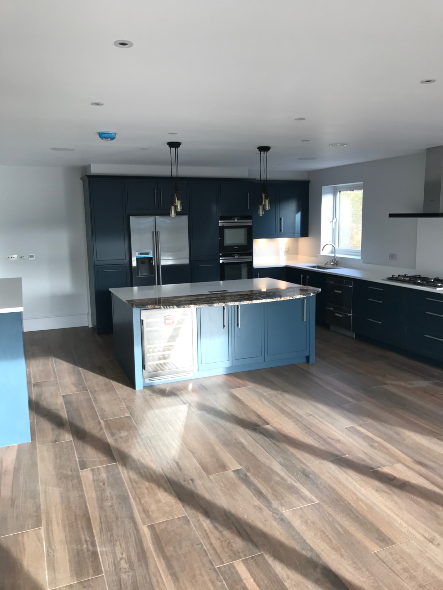 Nowoczesna, przestronna kuchnia z niebieskimi szafkami, wyspą z wbudowaną chłodziarką na wino i drewnopodobną podłogą z płytek, oświetlona wiszącymi lampami w stylu industrialnym.