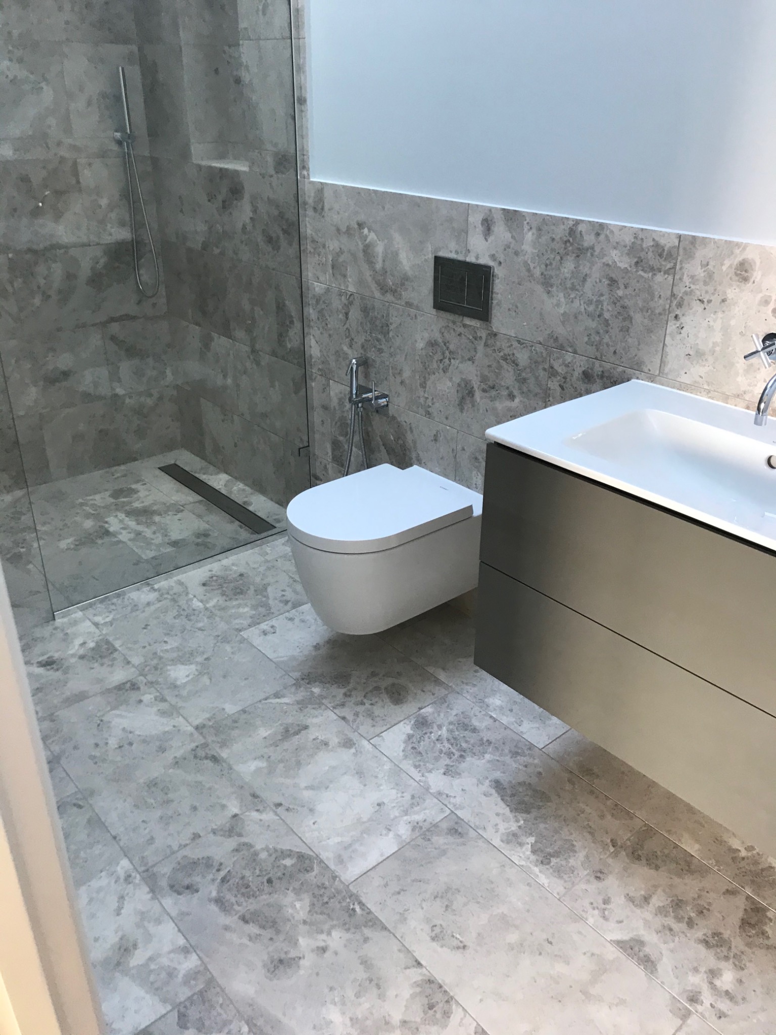 Nowoczesna łazienka wykończona szarym marmurem, z kabiną prysznicową typu walk-in, podwieszaną toaletą i szafką pod umywalką w kolorze grafitowym.