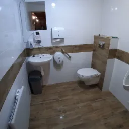 Nowoczesna toaleta publiczna z białymi i drewnopodobnymi płytkami, umywalką, pisuarami i toaletą wiszącą.