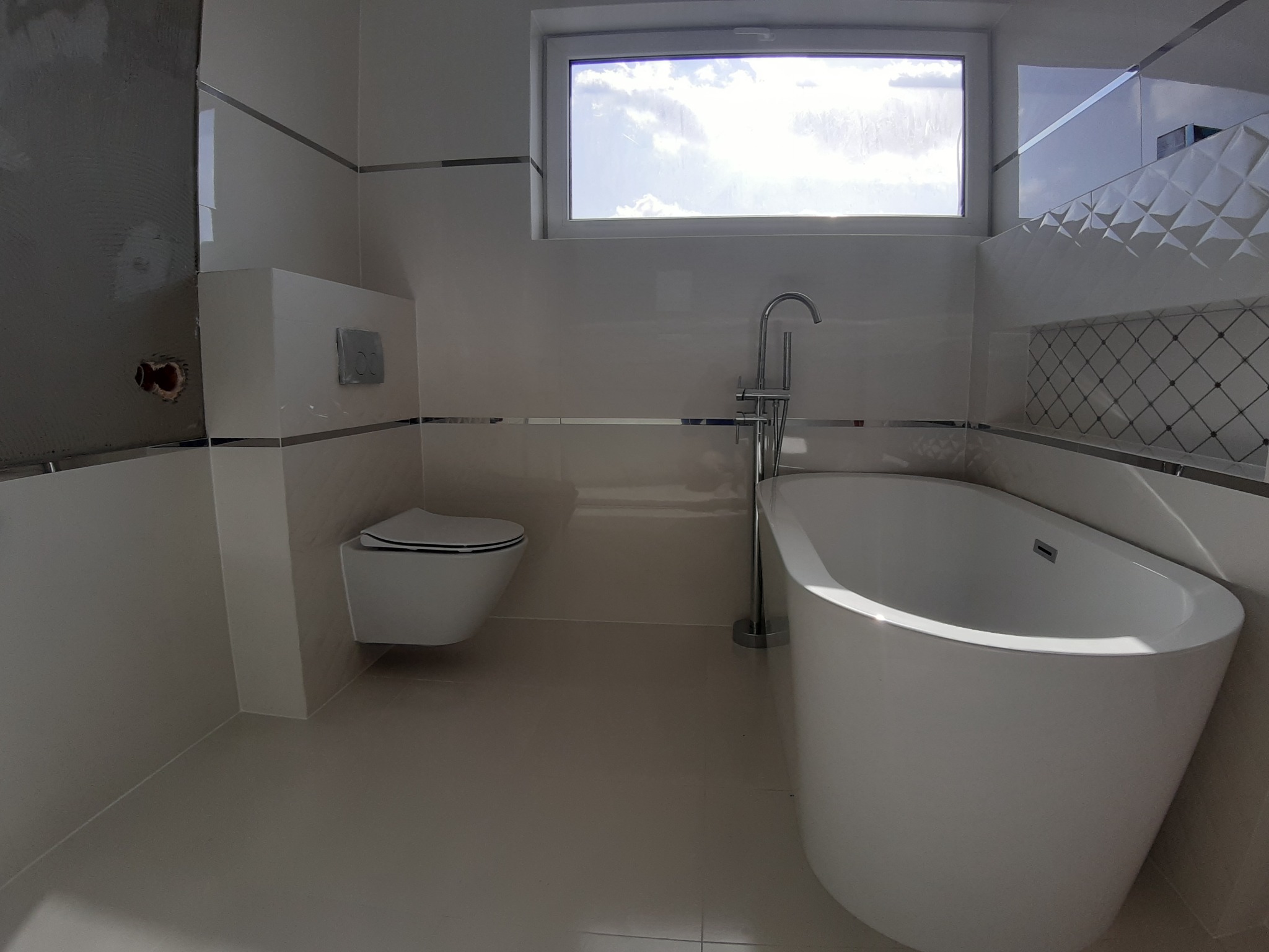 Nowoczesna łazienka z wolnostojącą wanną, podwieszaną toaletą i jasnymi płytkami, częściowo wykończona, z widocznym fragmentem ściany przygotowanej pod glazurę.