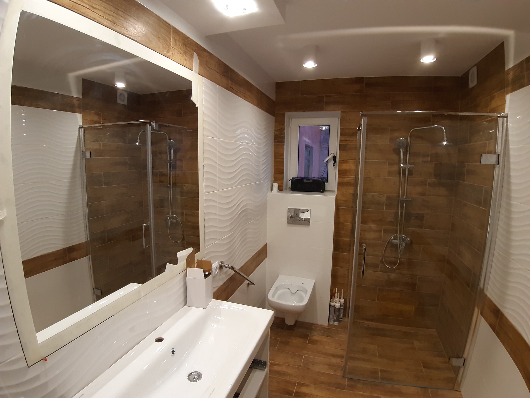 Nowoczesna łazienka z kabiną prysznicową typu walk-in, białymi płytkami z falistym wzorem, drewnopodobnymi płytkami podłogowymi i ściennymi, oraz białą umywalką z szafką, widoczne tuby z silikonem...