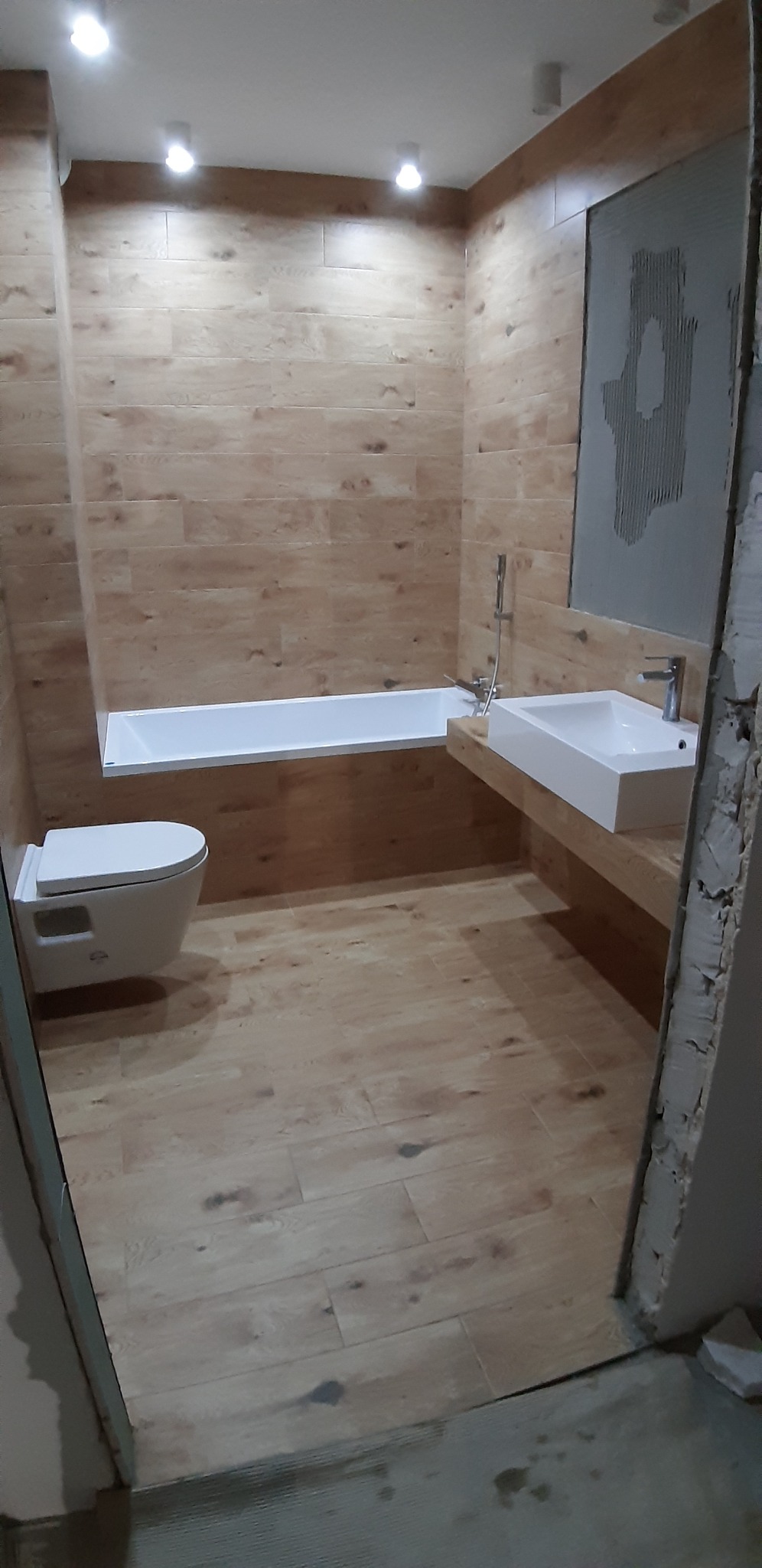 Nowoczesna łazienka z płytkami imitującymi drewno, białą armaturą i fragmentem ściany w trakcie układania płytek, widok z perspektywy wejścia.