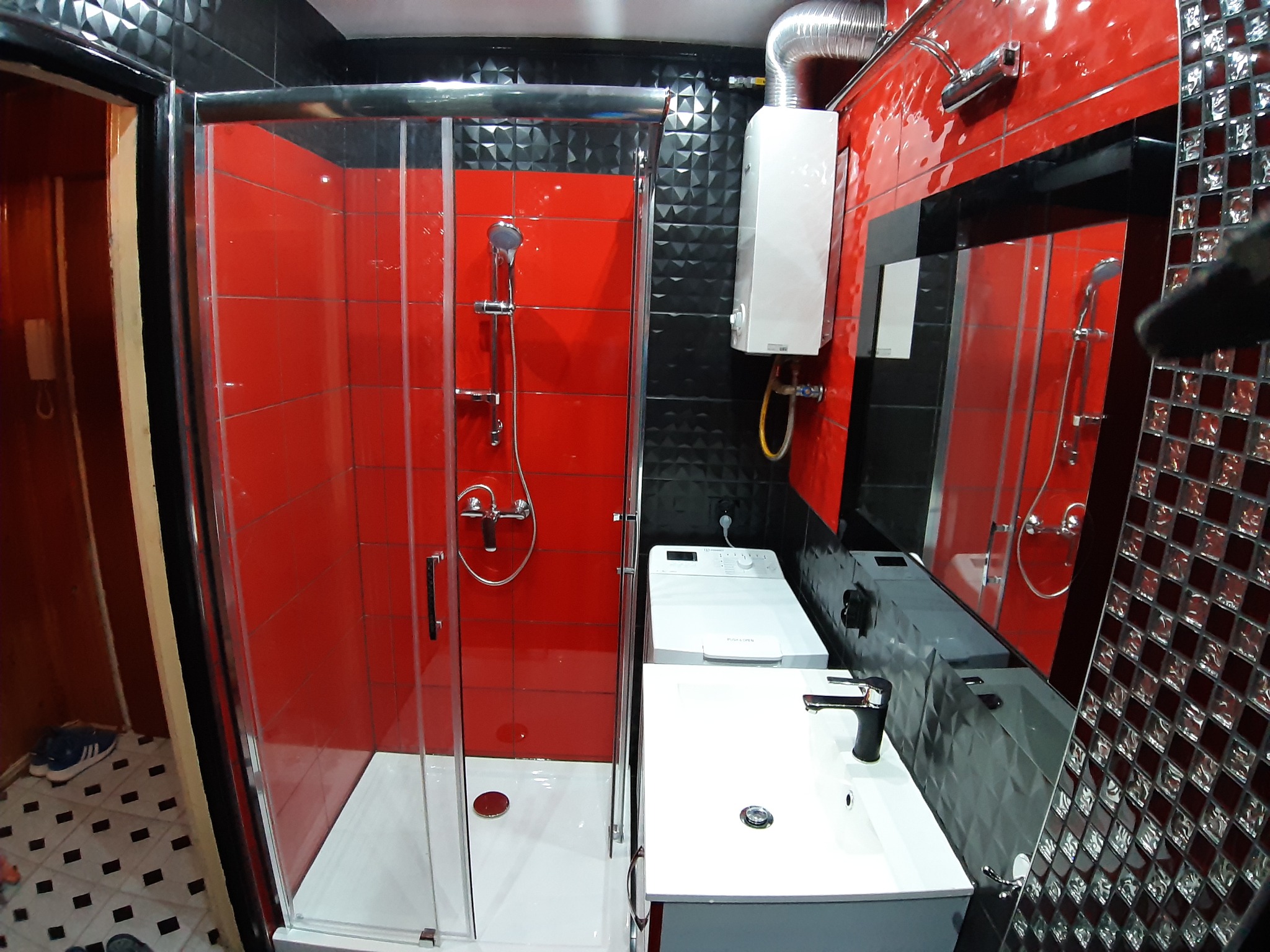 Nowoczesna łazienka w Ożarowie po remoncie: czerwone płytki w kabinie prysznicowej kontrastują z czarnymi, fasetowanymi płytkami na ścianach, biała umywalka i pralka, widoczny bojler.