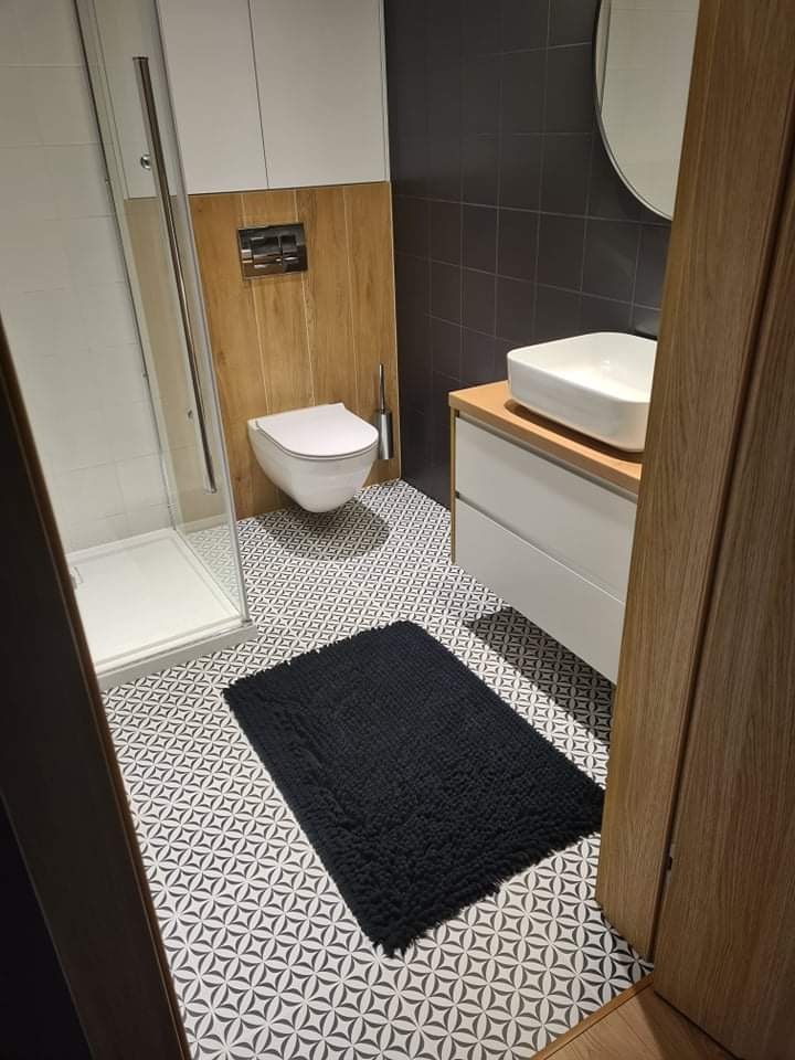 Nowoczesna łazienka z kabiną prysznicową, podwieszaną toaletą, umywalką nablatową i czarnym dywanikiem na wzorzystej podłodze.
