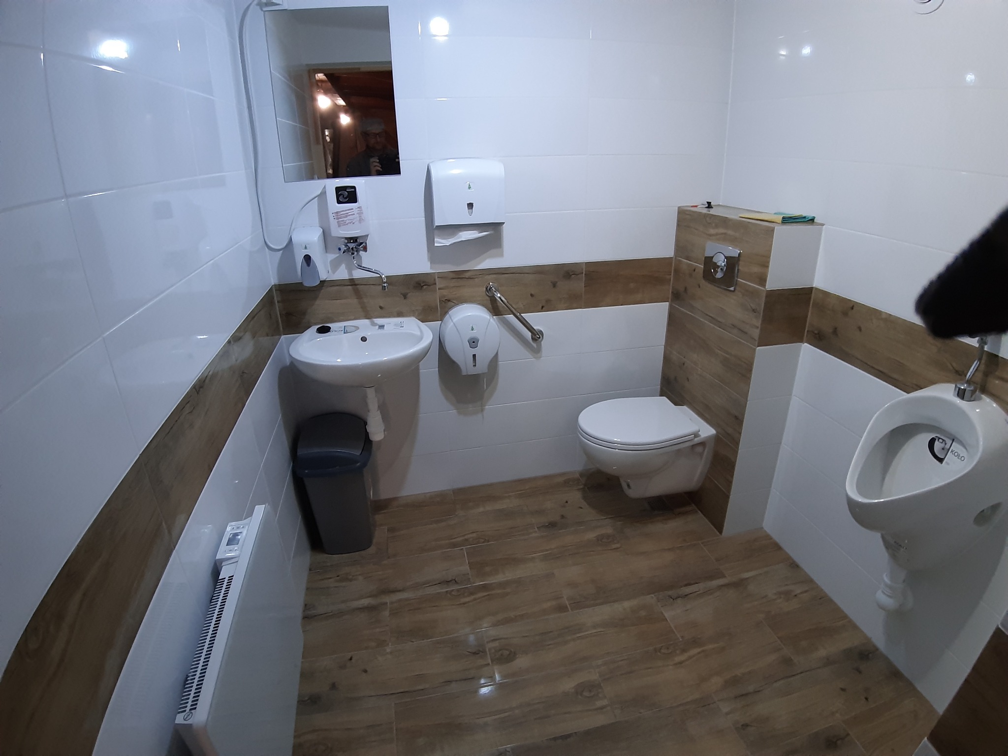 Nowoczesna toaleta publiczna z białymi i drewnopodobnymi płytkami, umywalką, pisuarami i toaletą wiszącą.