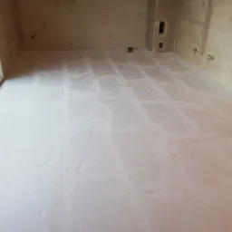 Surowy stan posadzki w pomieszczeniu z widocznymi śladami po płytkach, przygotowana do dalszych prac wykończeniowych, ściany w stanie deweloperskim z widocznymi puszkami elektrycznymi.