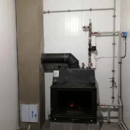 Wnętrze kotłowni z zainstalowanym piecem na paliwo stałe, widoczne rury instalacji grzewczej, komin w trakcie wykończenia, piec ustawiony na betonowych bloczkach