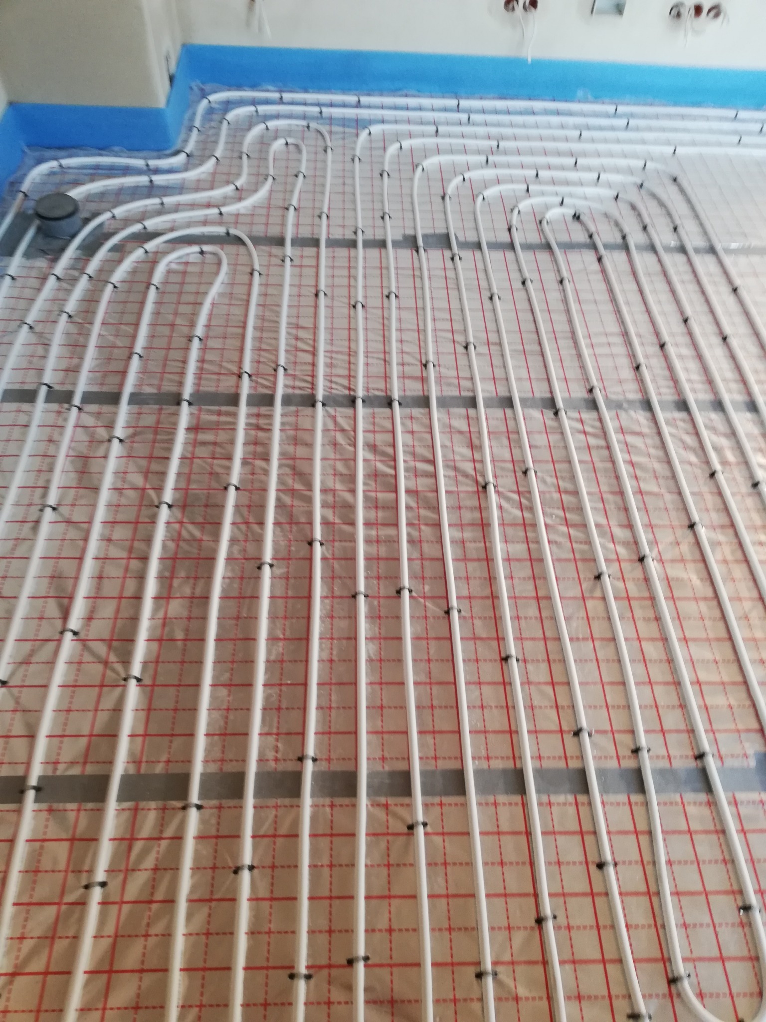 Białe rury ogrzewania podłogowego ułożone na folii z czerwoną kratką, przymocowane czarnymi klipsami, niebieska folia zabezpieczająca ściany wzdłuż krawędzi pomieszczenia.