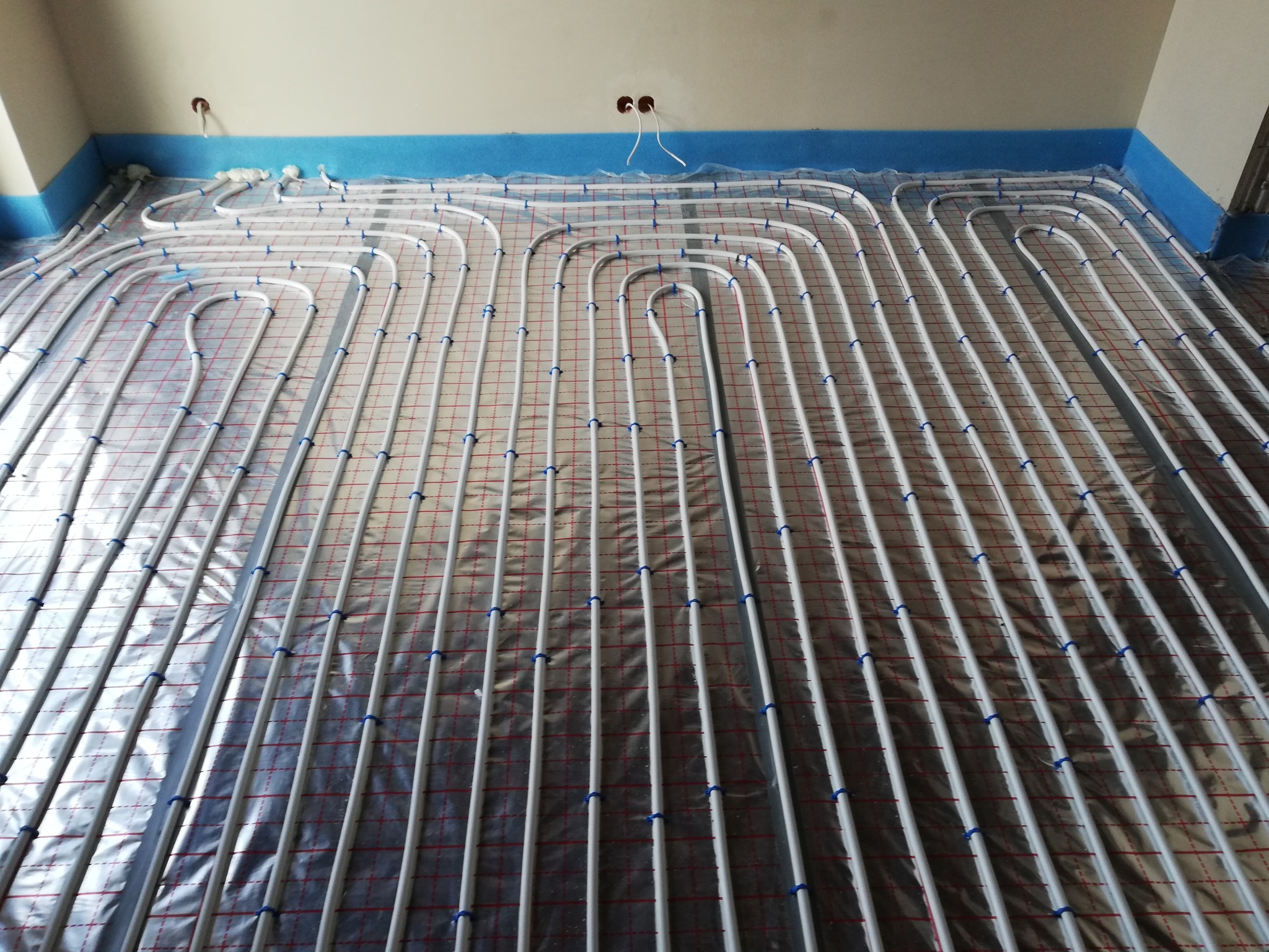 Rozłożone rury ogrzewania podłogowego na folii izolacyjnej z siatką, zabezpieczone niebieską taśmą przy ścianach, widoczne wystające kable elektryczne.