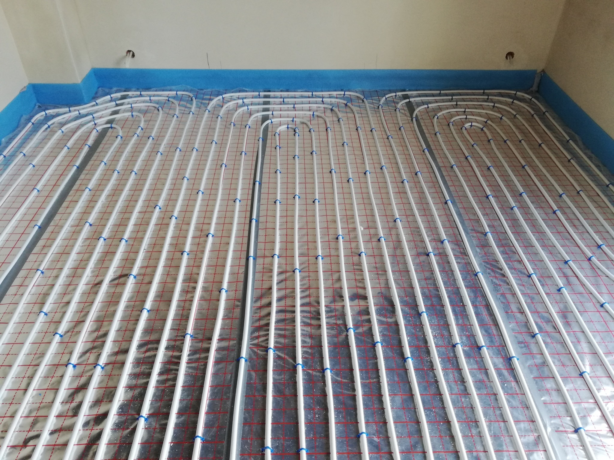 Białe rury ogrzewania podłogowego, przymocowane niebieskimi klipsami do folii z czerwoną siatką, ułożone na podłodze zabezpieczonej niebieską pianką przy ścianach.