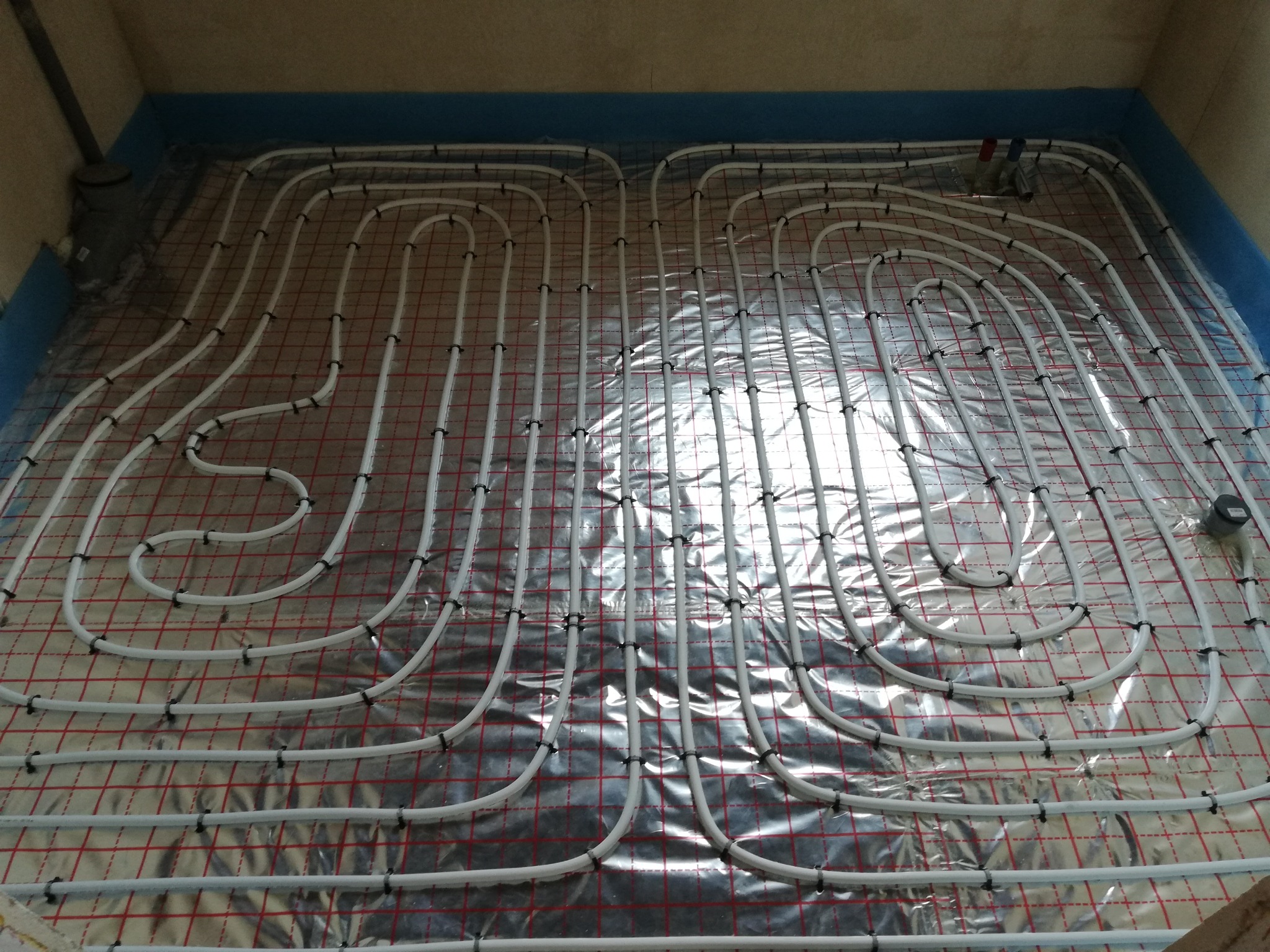 Rozłożone białe rury ogrzewania podłogowego na srebrnej folii z czerwoną kratką, przymocowane czarnymi klipsami, widok z góry na etapie instalacji w pomieszczeniu z szarymi ścianami.