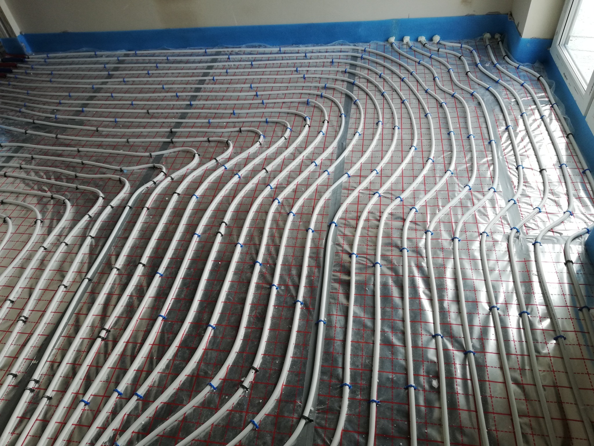 Rozłożone rury ogrzewania podłogowego na folii z siatką, przymocowane niebieskimi uchwytami, widok z góry, instalacja w trakcie realizacji.