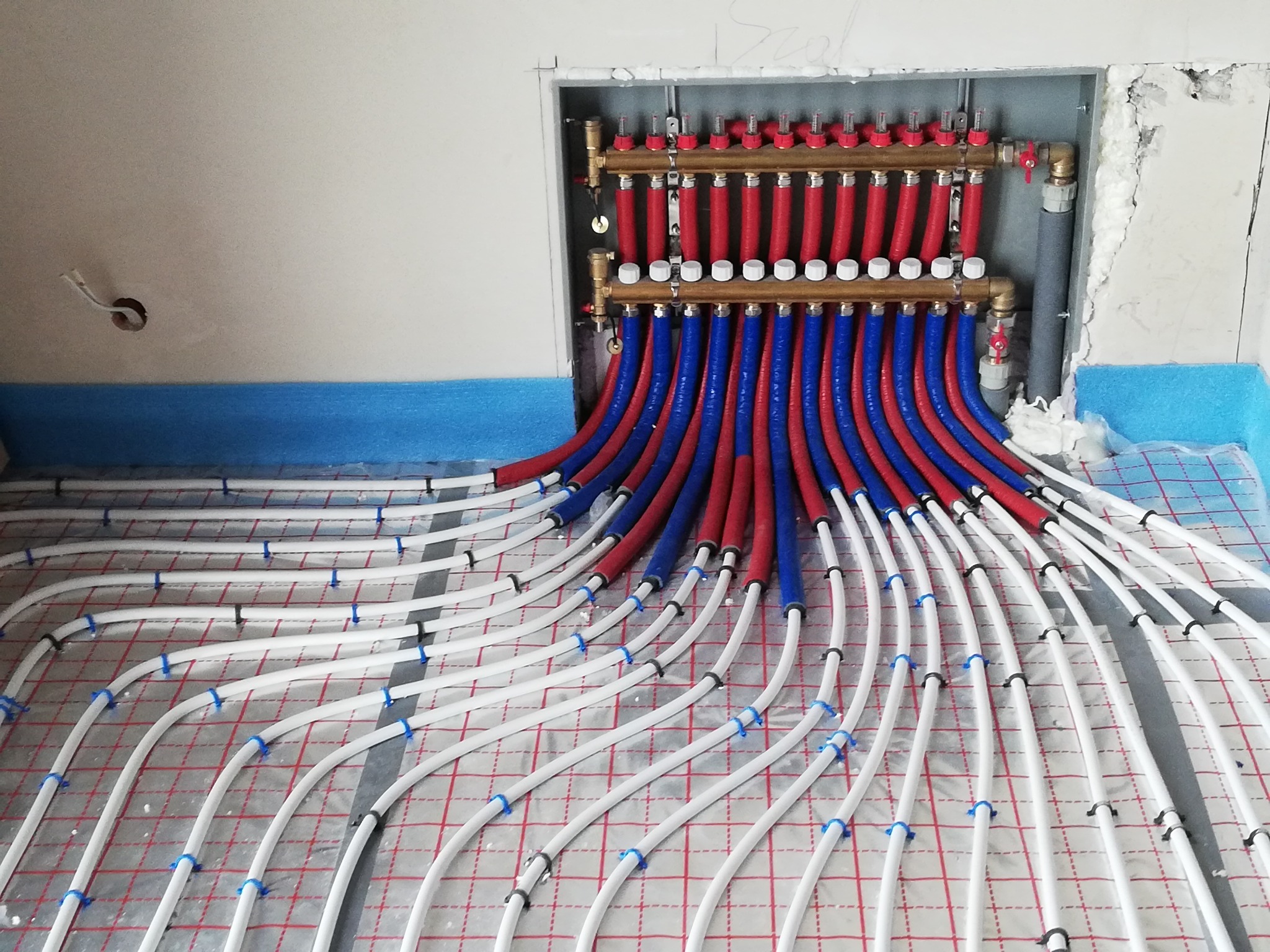 Rozdzielacz ogrzewania podłogowego z rurami w kolorach czerwonym i niebieskim, zamocowanymi do podłoża z siatką, widok z góry, w tle ściana w trakcie wykończenia.