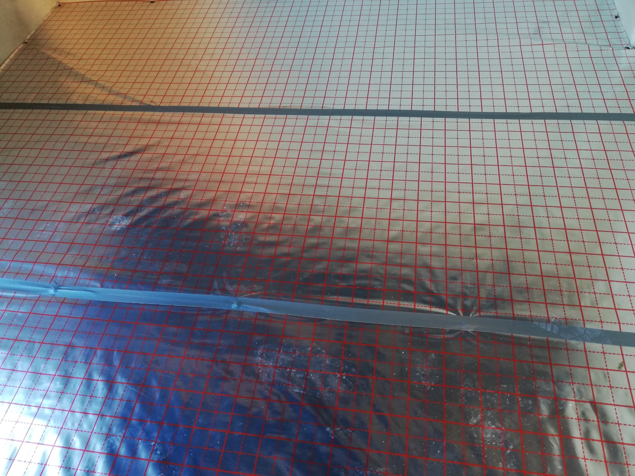 Folia aluminiowa z czerwoną siatką i szarą taśmą na podłodze, przygotowana pod instalację ogrzewania podłogowego.