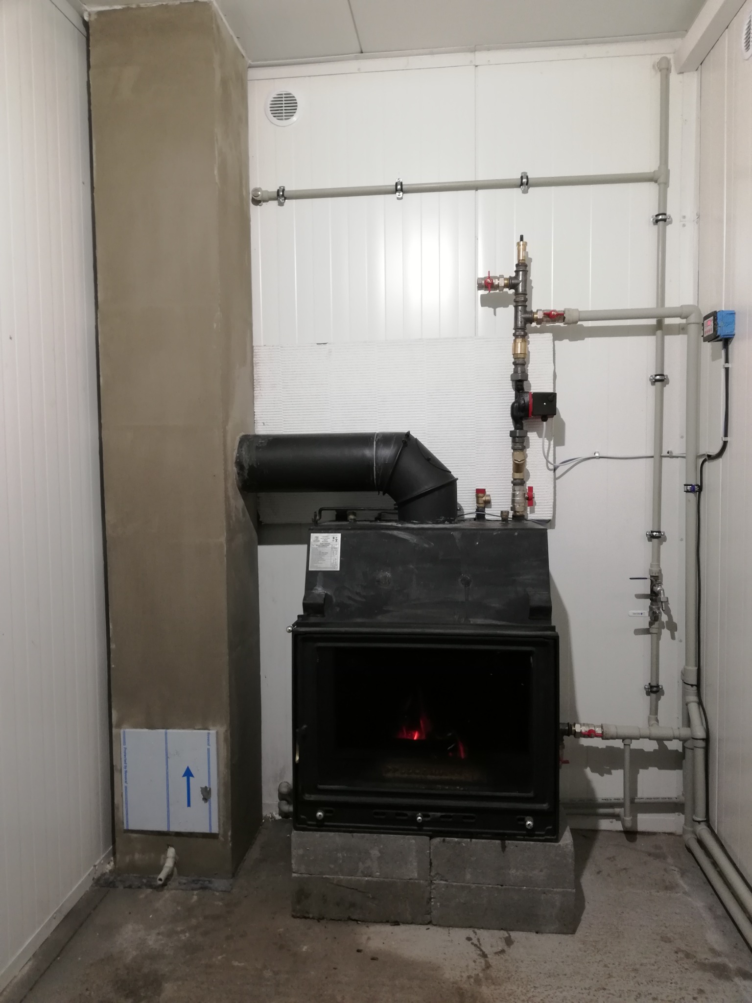 Wnętrze kotłowni z zainstalowanym piecem na paliwo stałe, widoczne rury instalacji grzewczej, komin w trakcie wykończenia, piec ustawiony na betonowych bloczkach