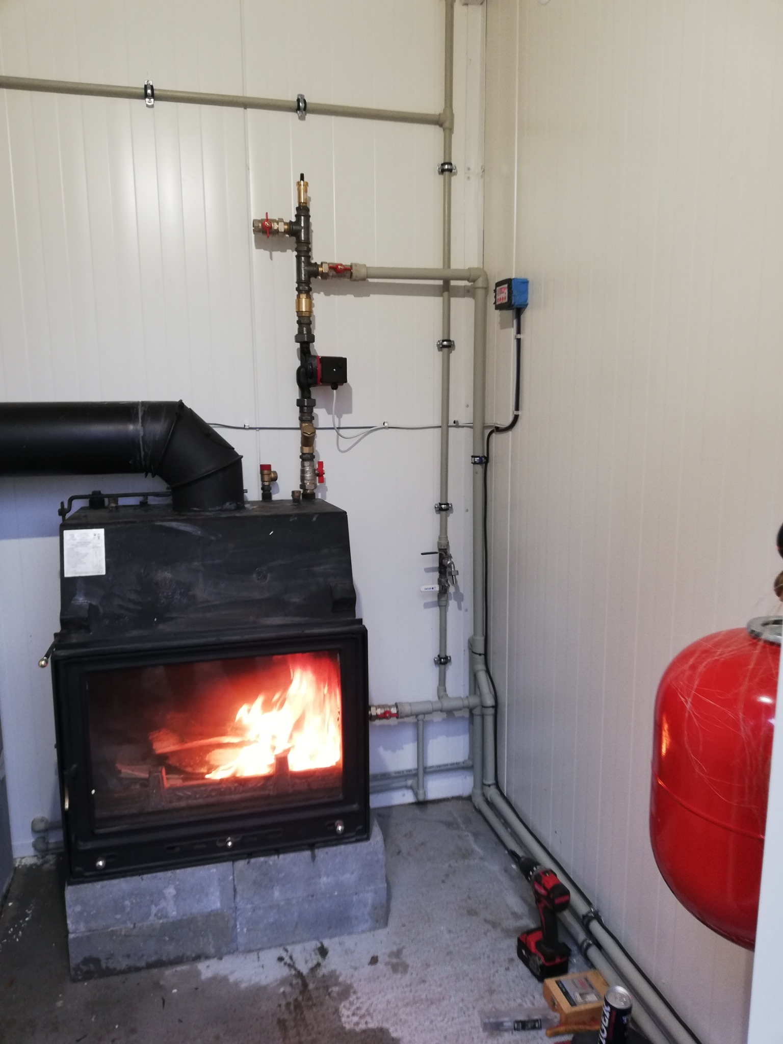 Czarny piec kominkowy z palącym się drewnem, ustawiony na betonowych bloczkach, z podłączonymi rurami i czerwonym zbiornikiem wyrównawczym. W tle białe panele ścienne i narzędzia na podłodze.