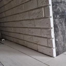 Fragment ściany wykończonej dekoracyjną cegłą w odcieniach bieli, sąsiadującej z płytami imitującymi czarny marmur, na podłodze jasne płytki imitujące drewno.