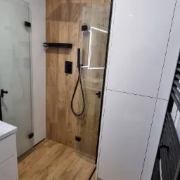 Nowoczesna kabina prysznicowa typu walk-in z czarną armaturą, drewnopodobne płytki na ścianie i podłodze, białe meble łazienkowe.