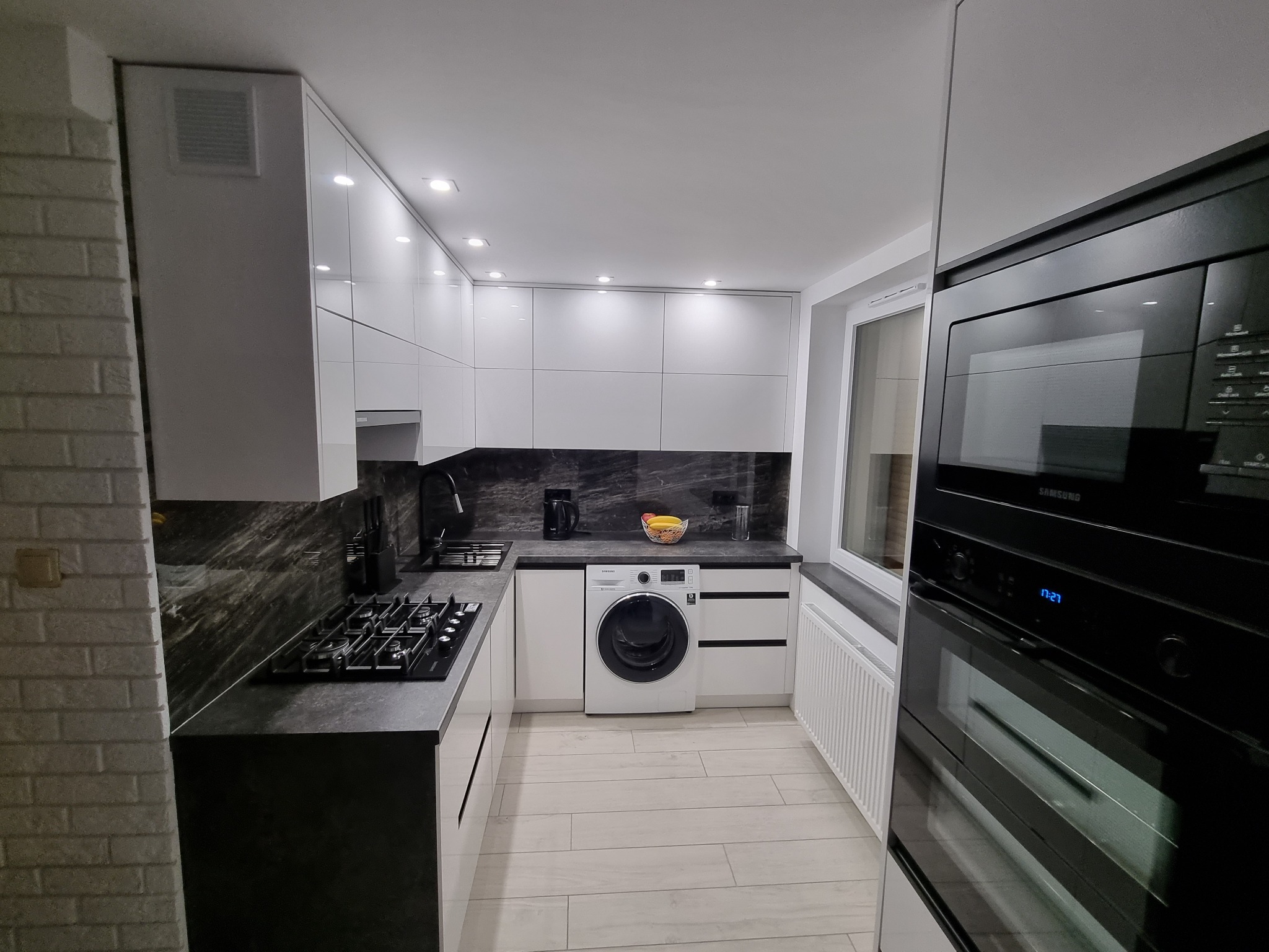 Nowoczesna kuchnia z białymi szafkami, blatem imitującym ciemny marmur, płytą gazową, piekarnikiem i pralką zabudowaną pod blatem.