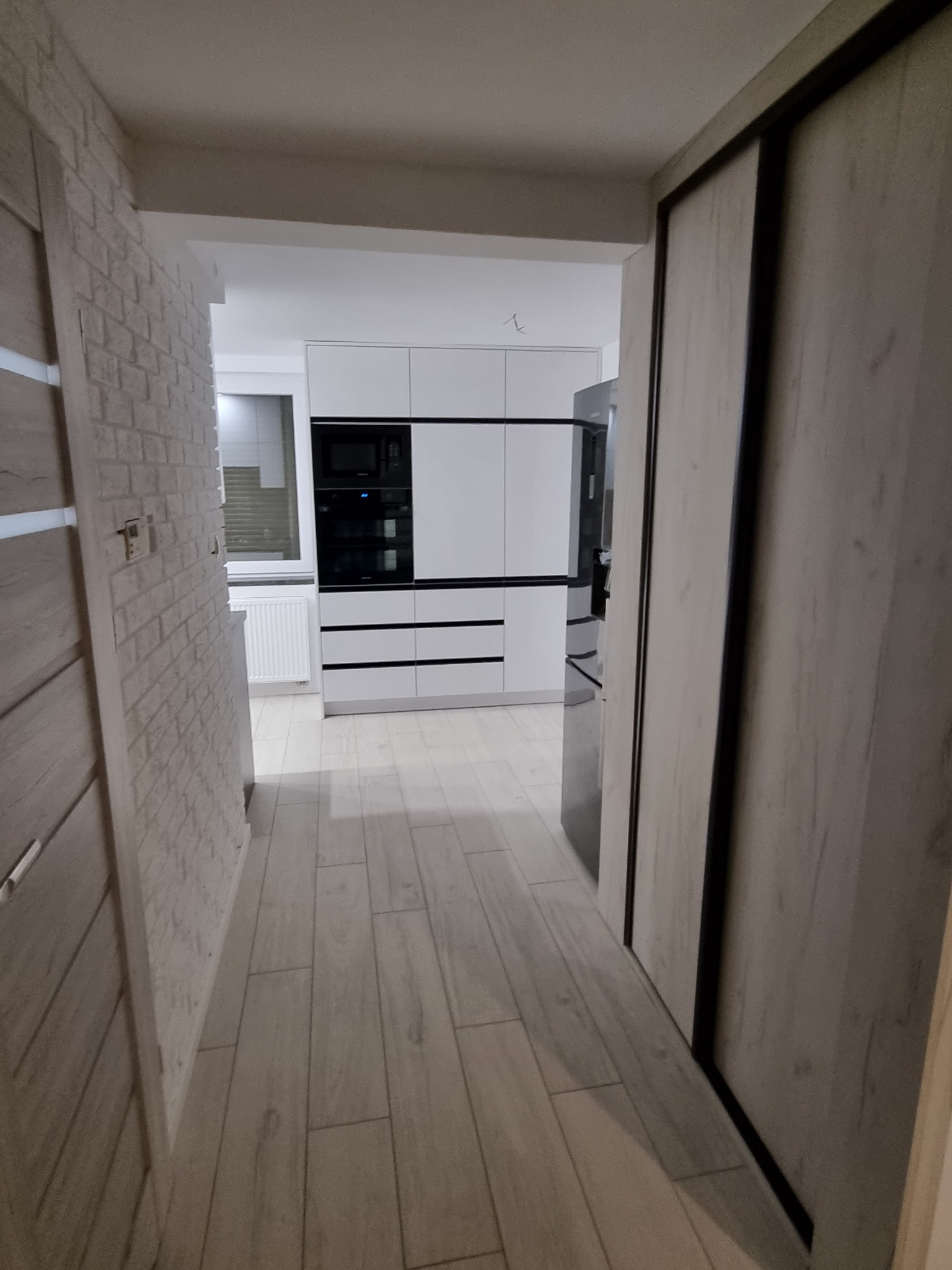Korytarz z widokiem na nowoczesną, białą kuchnię z czarnymi akcentami, jasną podłogą imitującą drewno i ścianą wykończoną cegłą dekoracyjną.