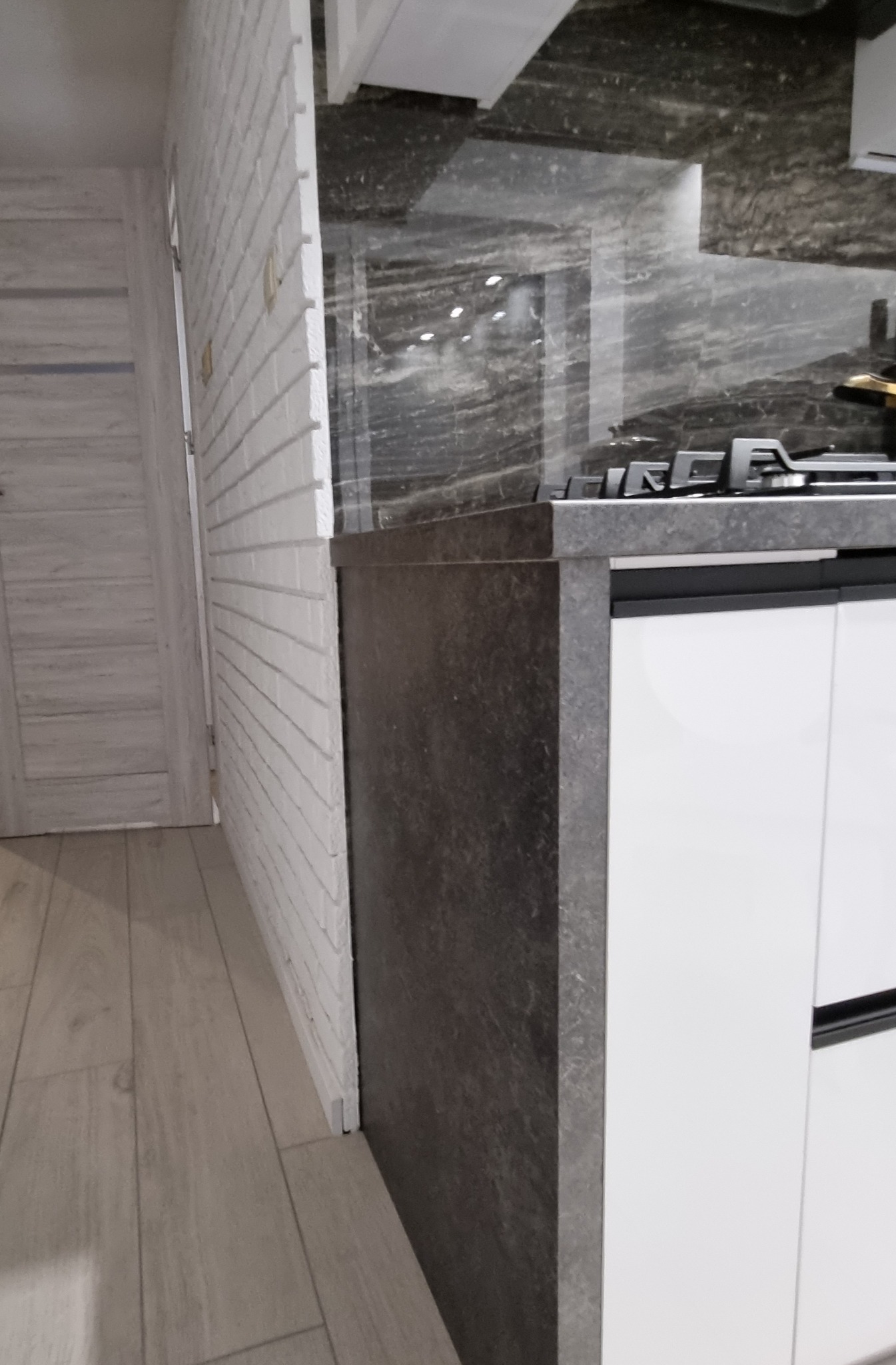 Fragment wykończonej kuchni: biała cegła na ścianie, szara marmurkowa płyta nad blatem, zabudowa kuchenna z białymi frontami i ciemnoszarym blatem, widoczna płyta gazowa.