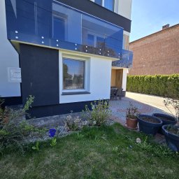 mfast - Nowoczesny dom z czarno-białą elewacją i przeszkloną balustradą balkonu z ciemnego szkła, mocowaną na srebrnych elementach. Przed nim zielony trawnik i brukowany podjazd.